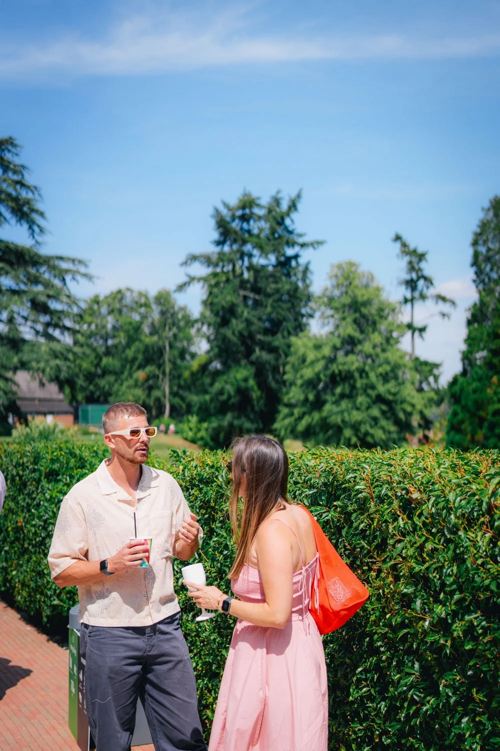A man and a woman conversing outdoors near a hedge, with trees and a blue sky in the background. The man is wearing sunglasses and holding a drink, and the woman is holding a cup and a reusable bag.