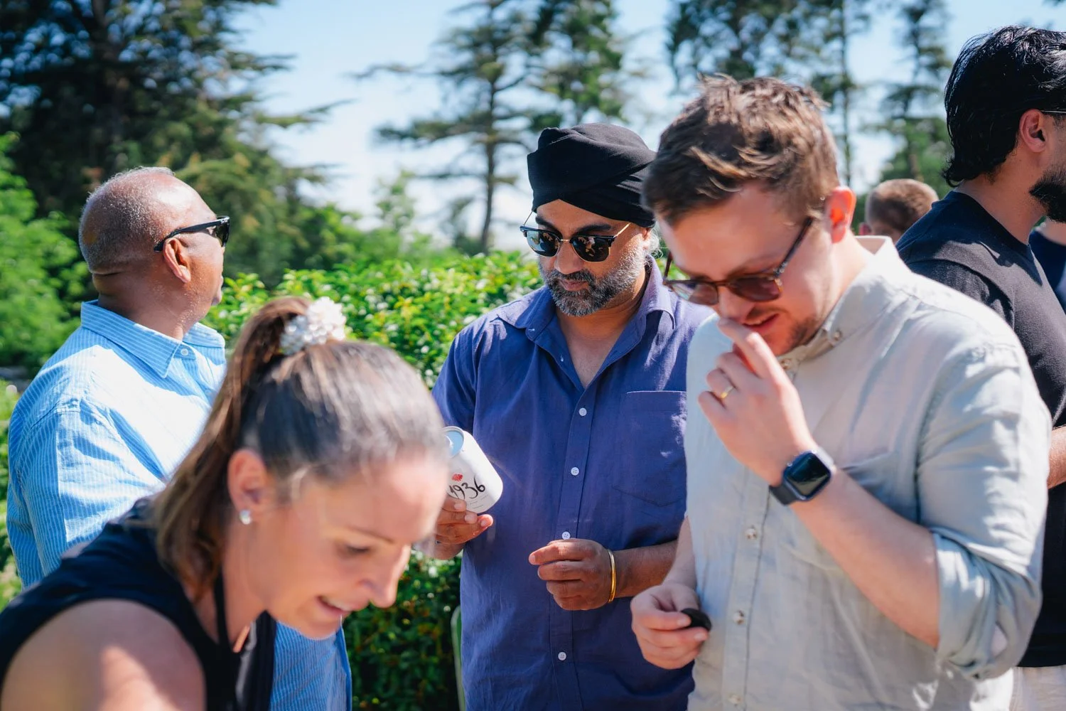 Group of people outdoors having a conversation, some wearing sunglasses, in a sunny setting with trees and green foliage in the background.
