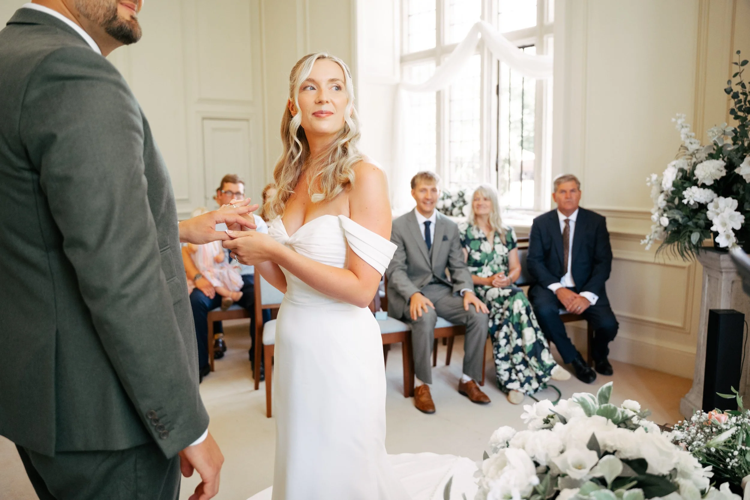 A bride with long blonde hair and a white dress holds hands with a man in a gray suit during a wedding ceremony in a bright room decorated with flowers.