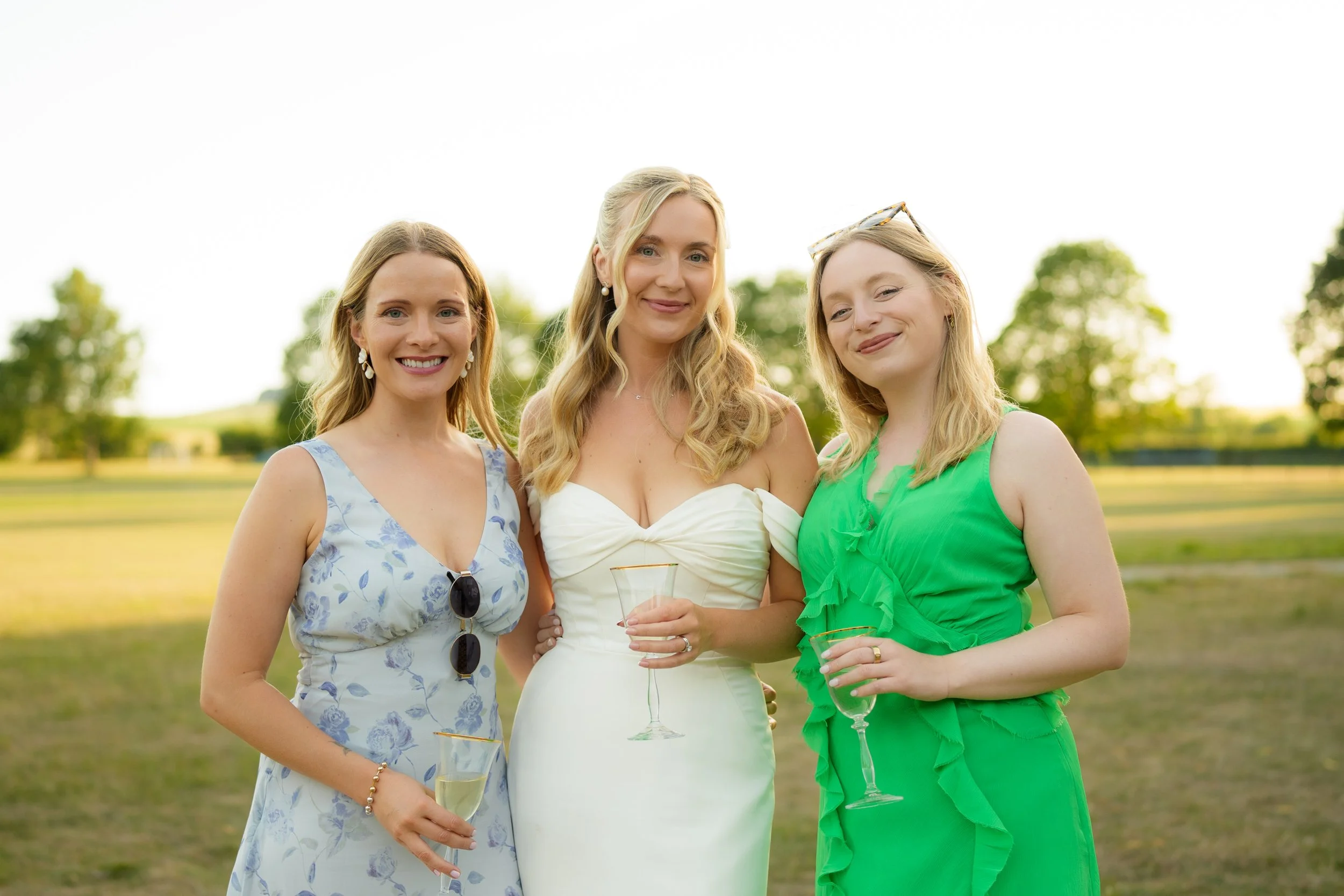 Three women dressed in summer attire holding glasses of wine outdoors with trees and a clear sky in the background.