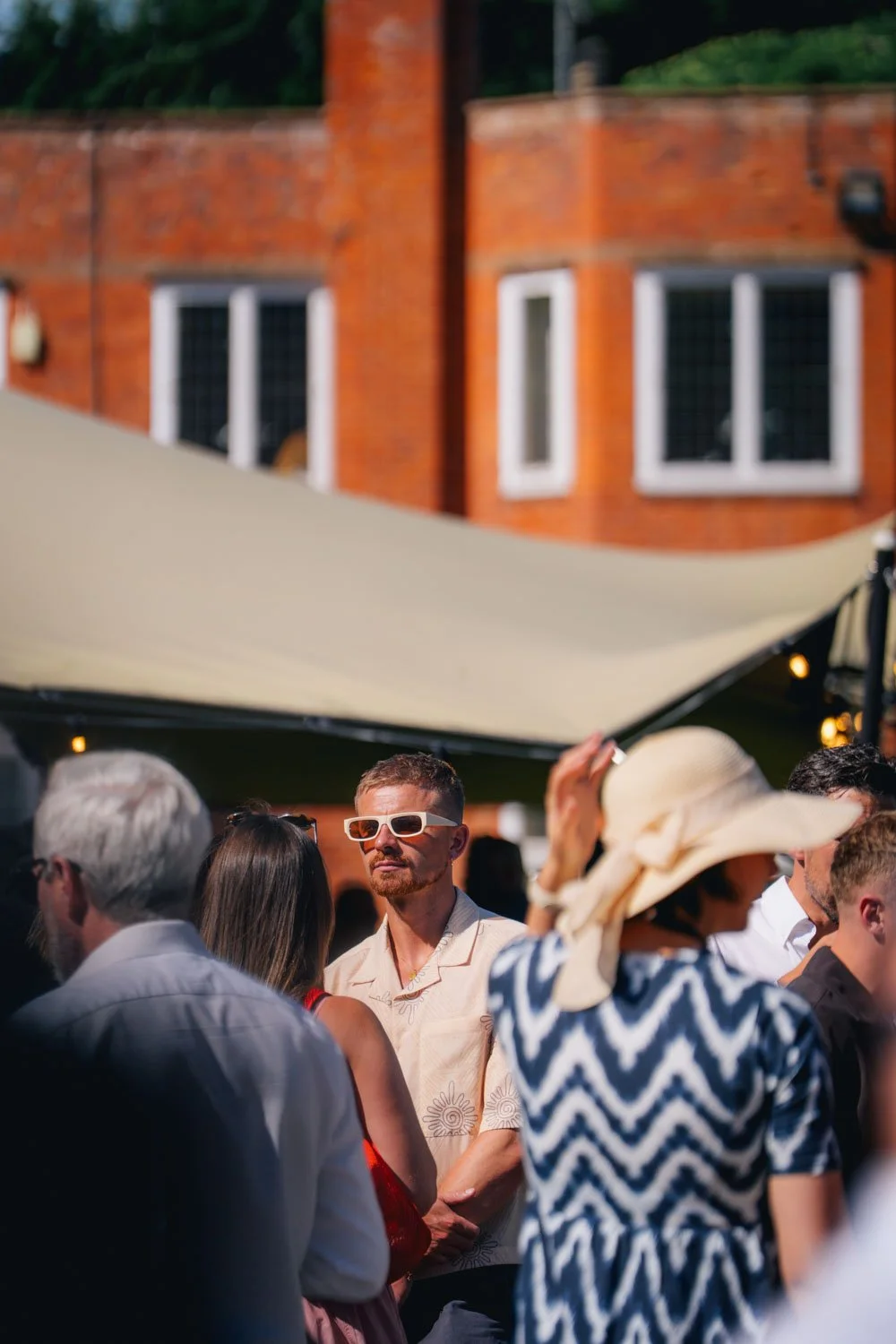 People gathered outdoors at a social event with a brick building and string lights in the background. The focus is on a man wearing white sunglasses and a short-sleeve patterned shirt.