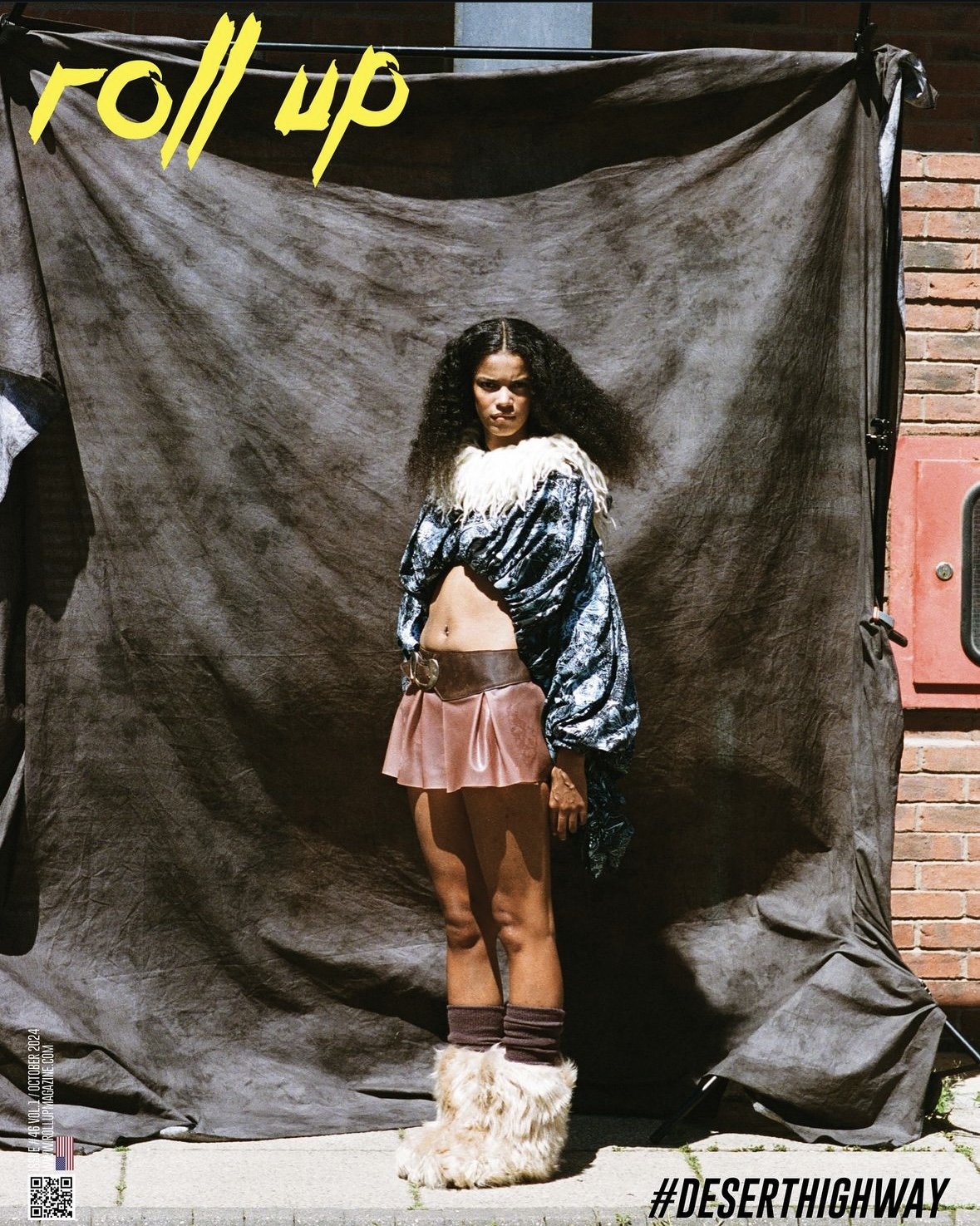 A woman standing in front of a gray backdrop, wearing a shiny patterned jacket, a short pink skirt, furry boots, and brown leg warmers, with a serious expression.