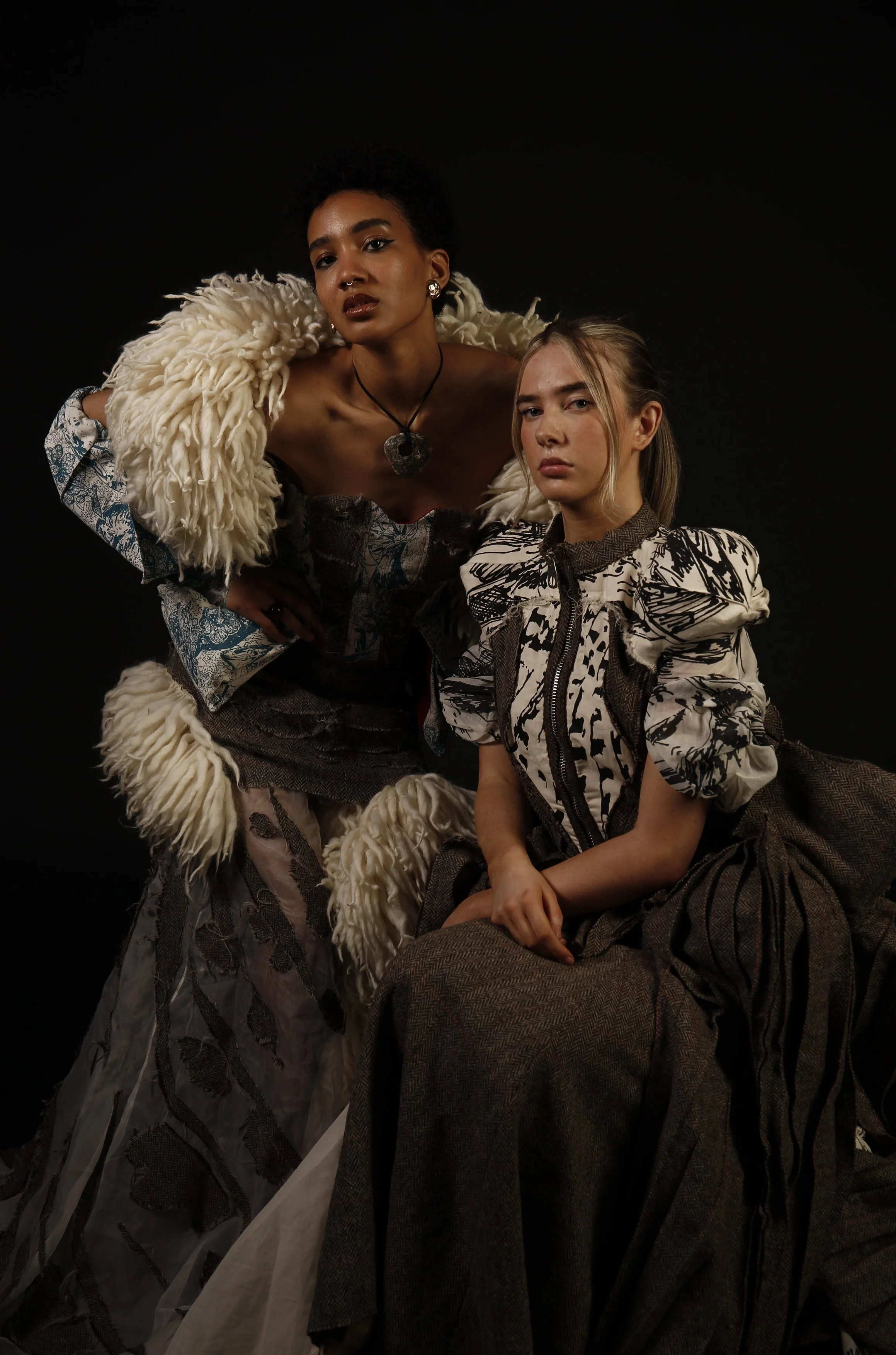 Two women posing in fashion outfits against a black background