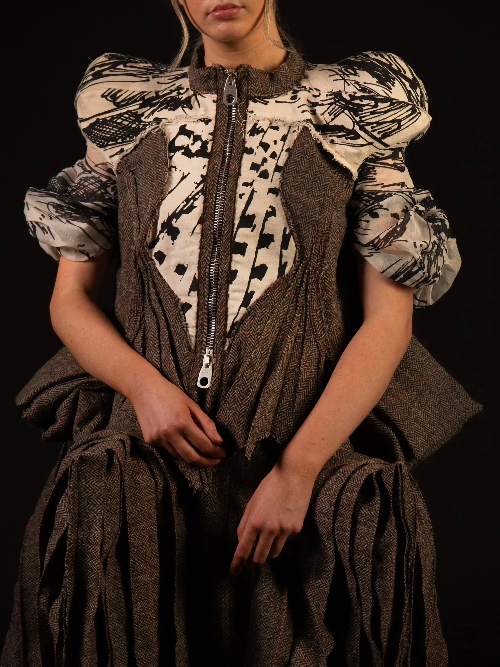 A person wearing a layered outfit with a patterned shirt, a brown patchwork vest, and a matching skirt against a black background.