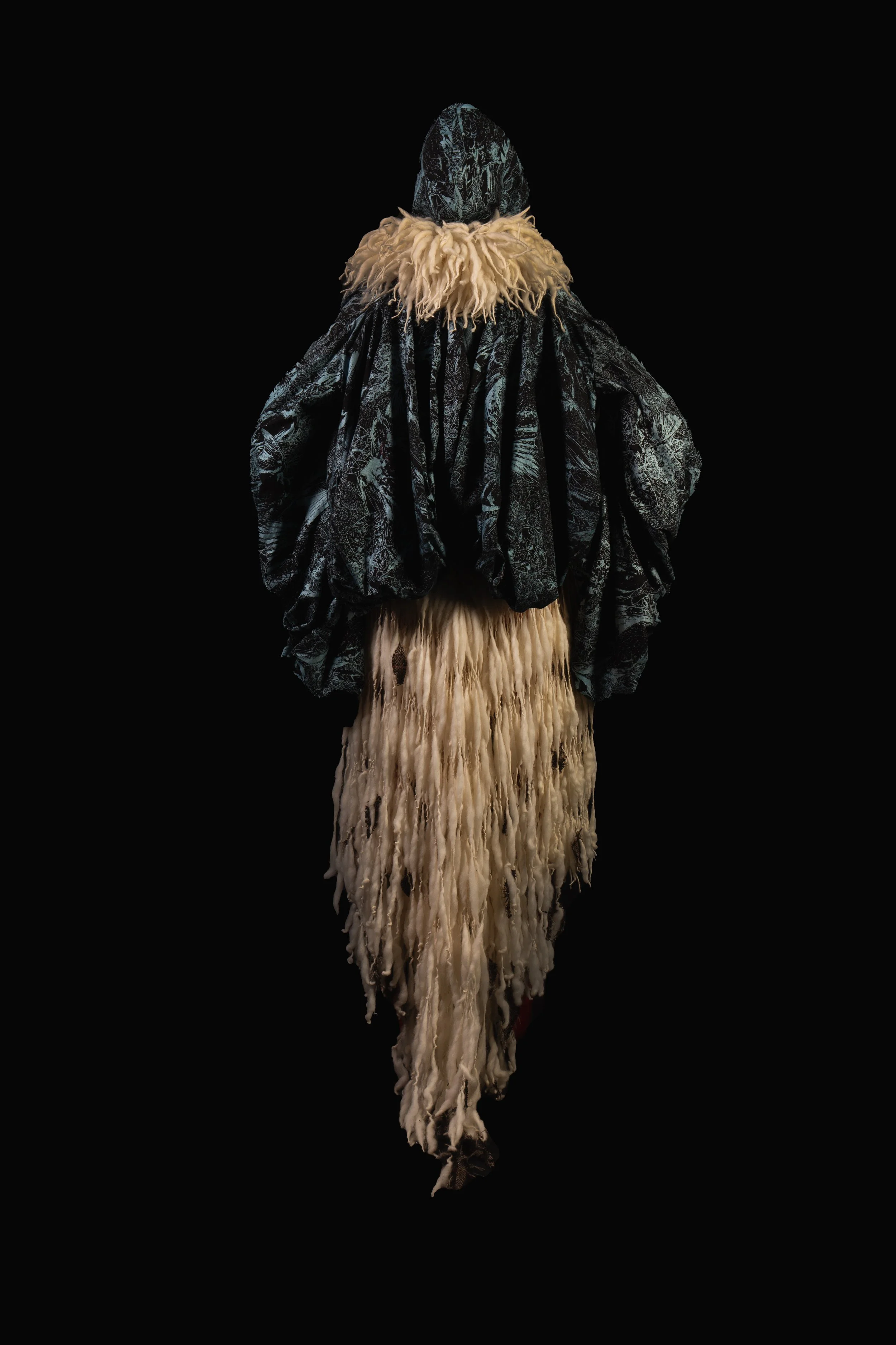 Fashion mannequin dressed in a black patterned cloak, with a beige furry collar and a long beige fringed skirt, stood against a black background.