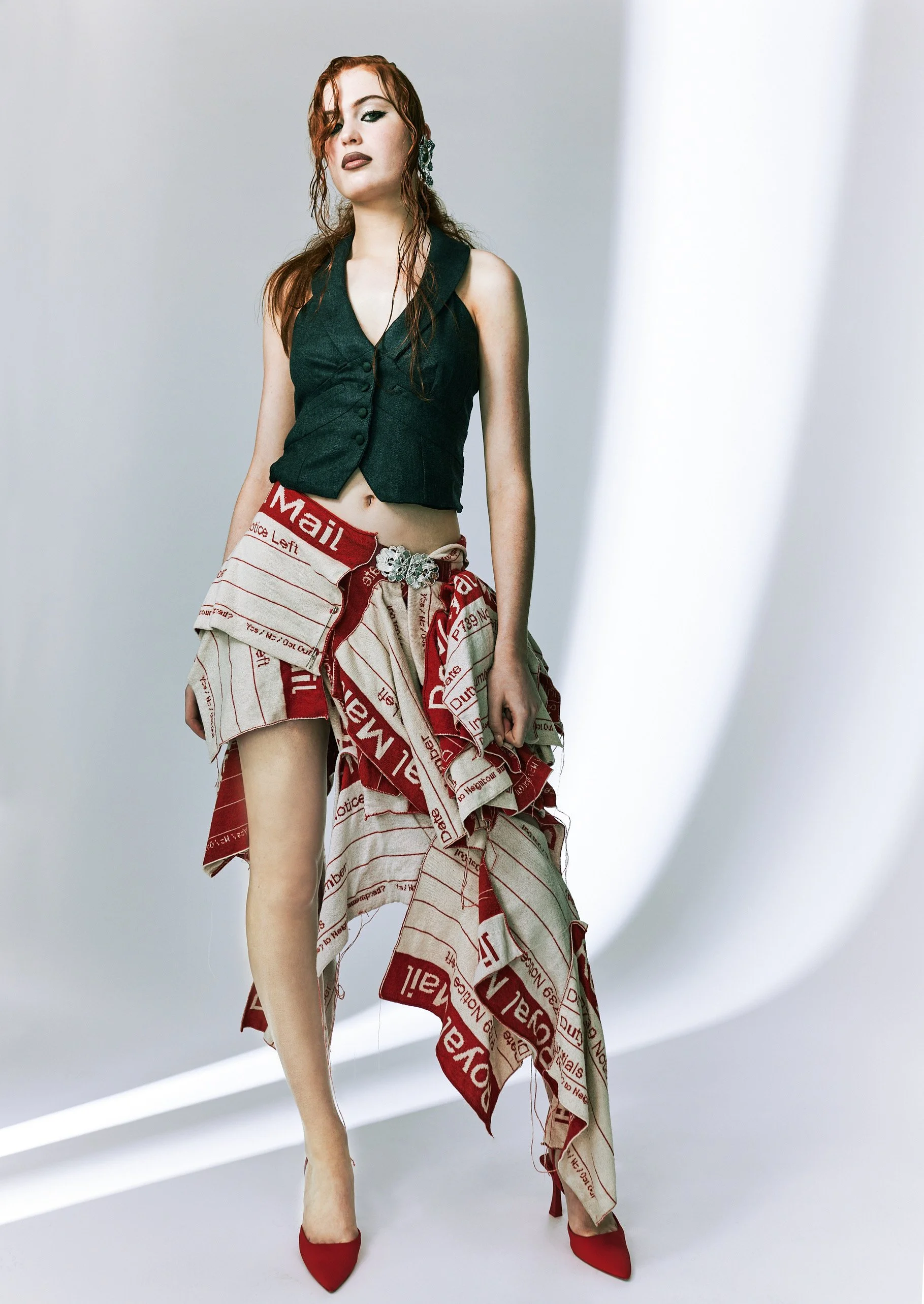 A woman with red hair and fair skin poses in a studio against a white background. She wears a black sleeveless vest, a paper print skirt with red and beige text and patterns, and red high heels. The skirt is styled with layered fabric, and she access