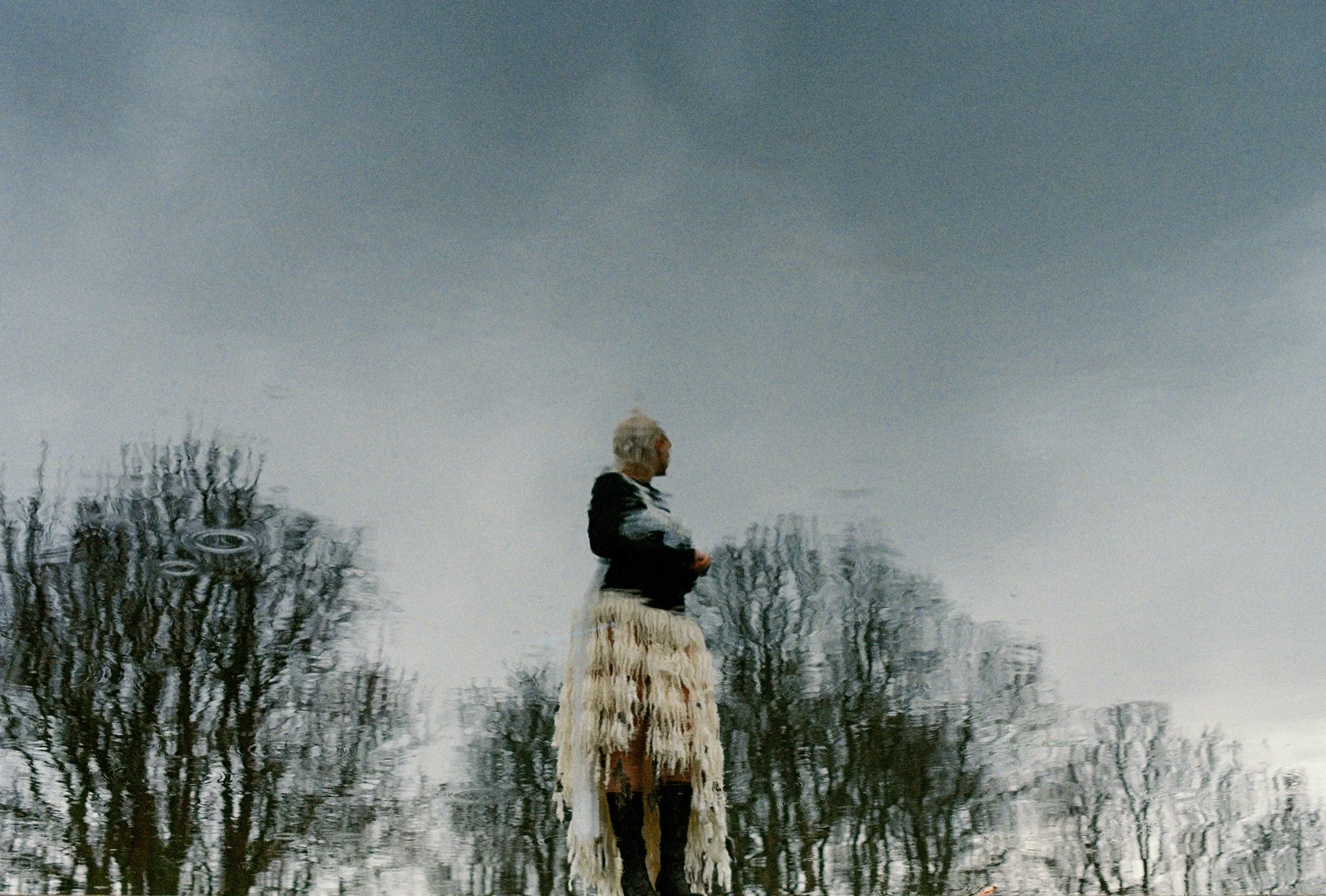A reflection of a person standing outdoors, wearing a long, fringed skirt and a black top, with trees and cloudy sky in the background.