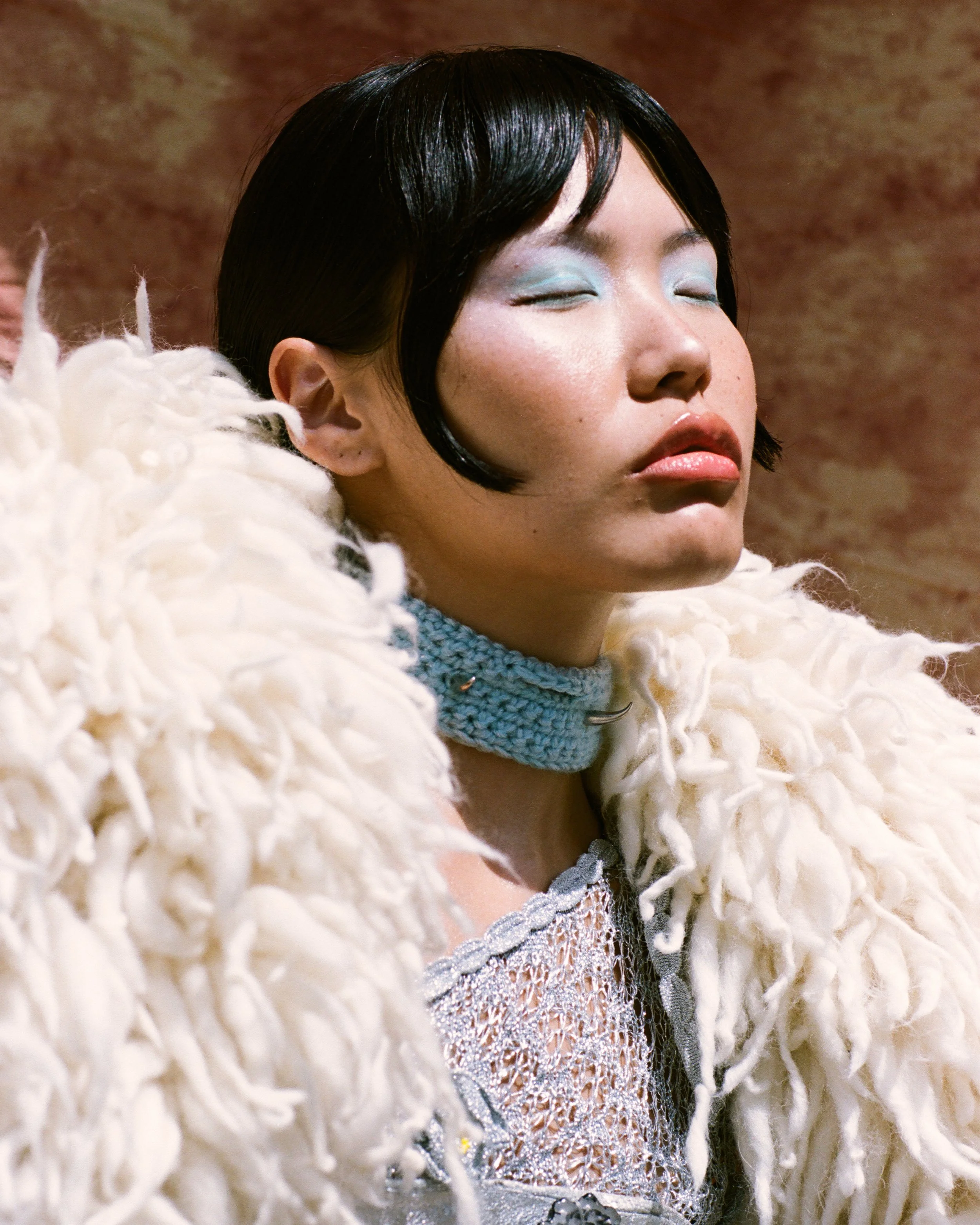 A woman with short black hair and closed eyes wearing a fluffy white coat and a light blue crochet choker neckband.