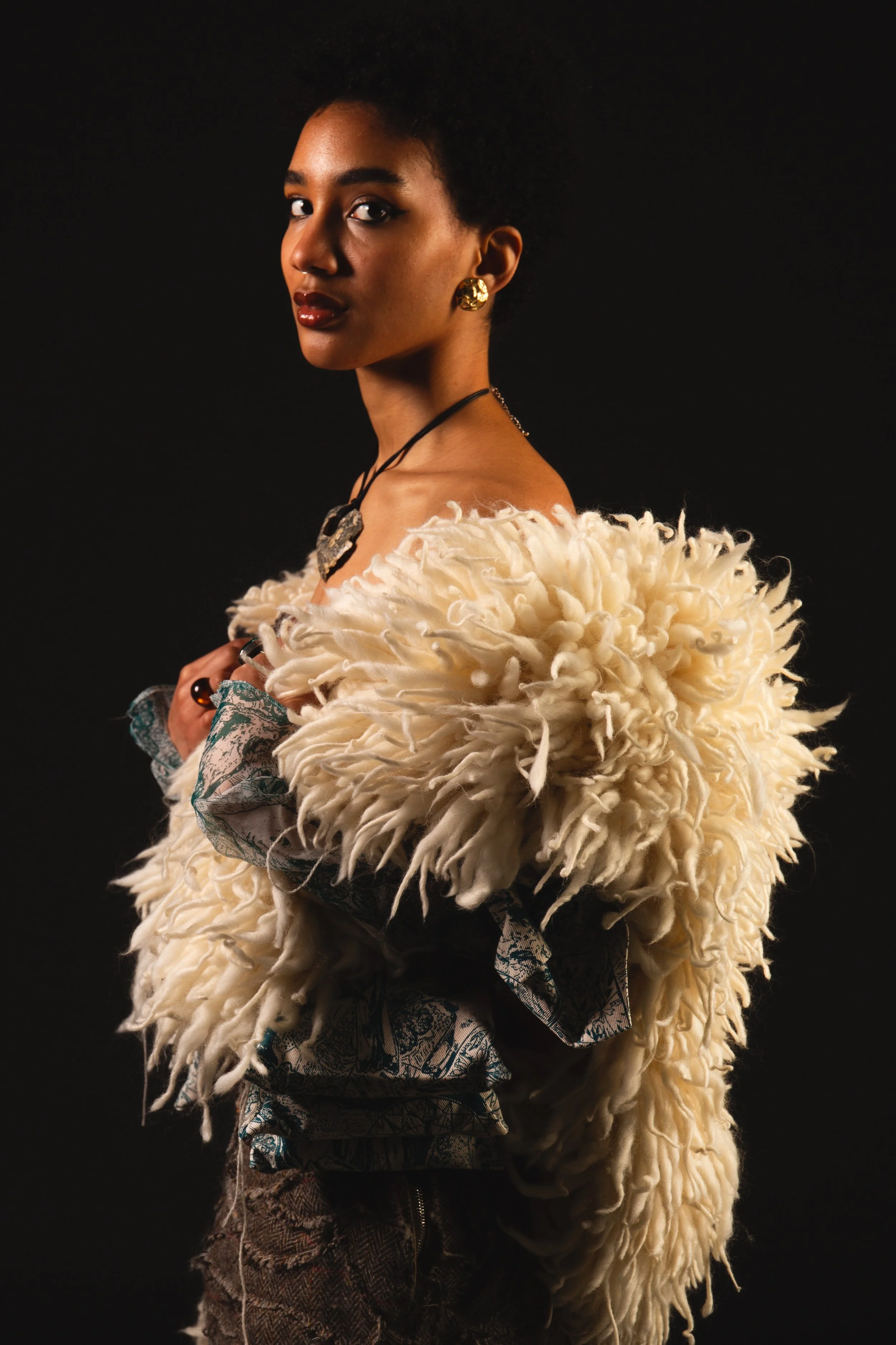 A woman with dark curly hair wearing gold earrings and a black necklace, dressed in a fluffy cream-colored garment layered over a patterned shirt, standing against a black background.