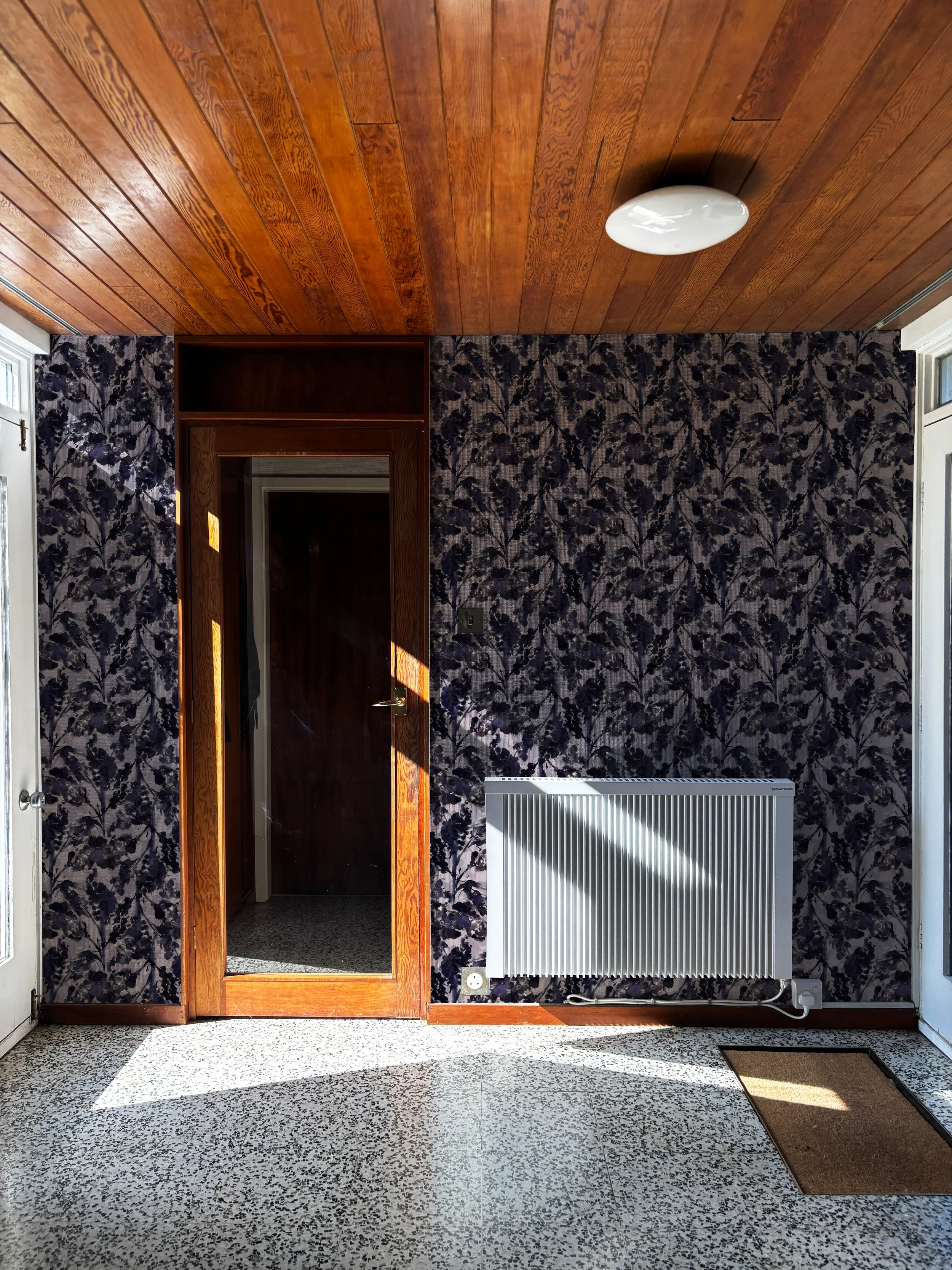 A room with a wooden ceiling, floral wallpaper, a white radiator, a mirror door, a brown doormat, and a small white electrical outlet.