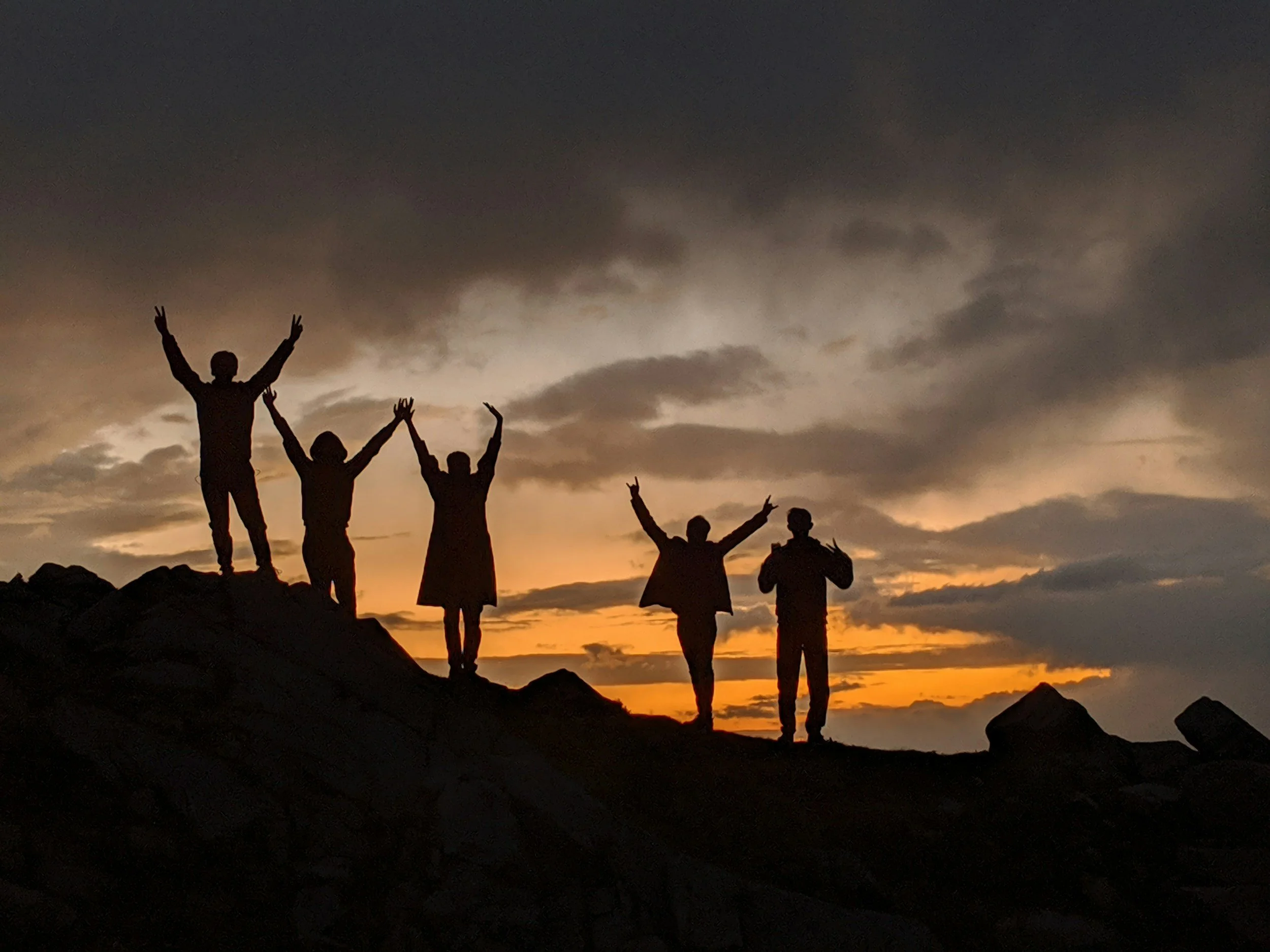 Silhouettes of five people standing on rocks during sunset, with five raising their arms in celebration against a cloudy sky.