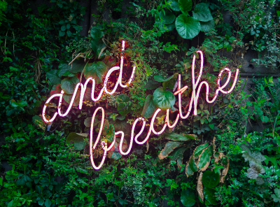 Neon sign reading 'and breathe' against a background of green foliage.