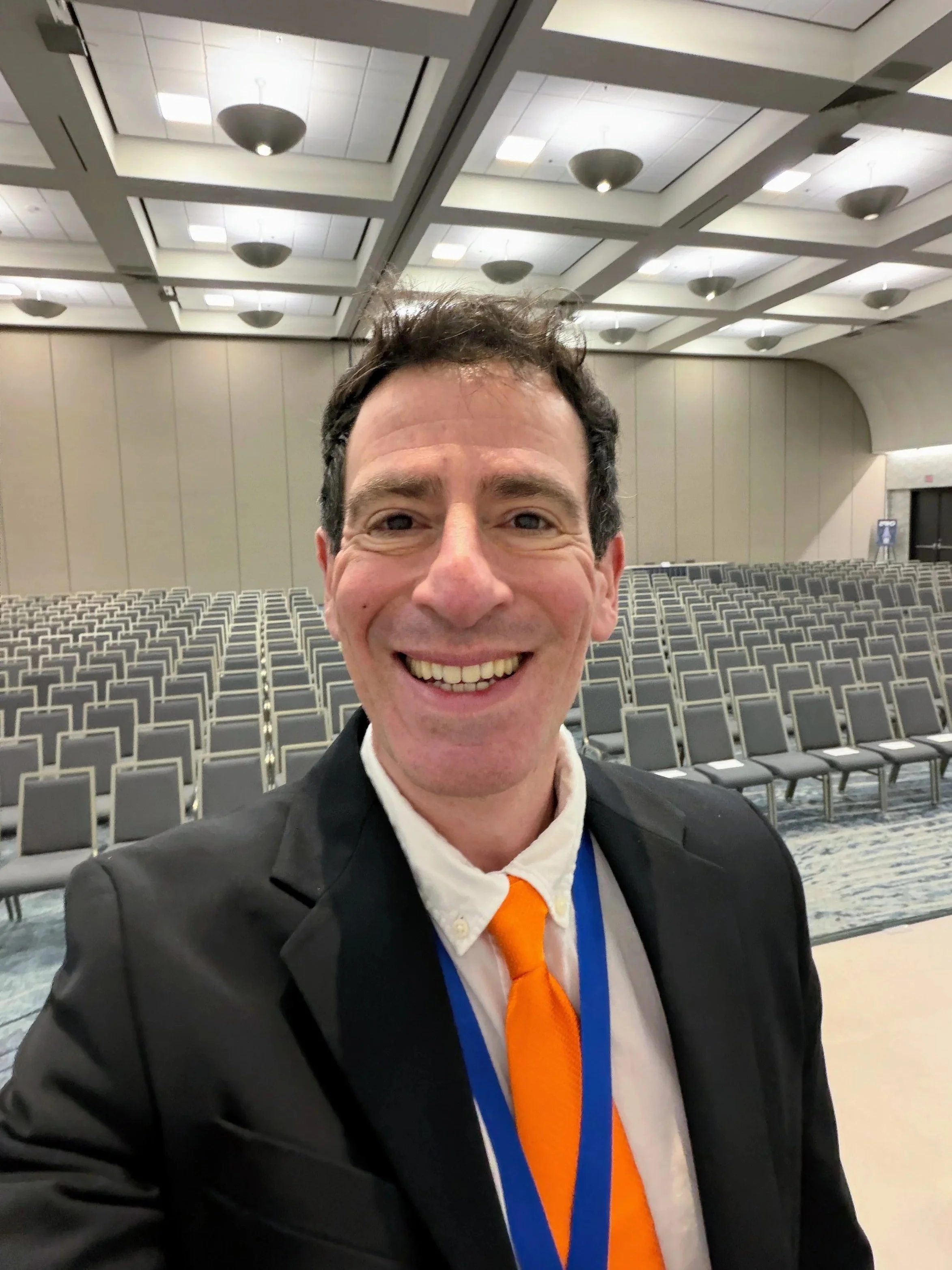 Dr. Gregory Charlop, MD, DipABLM, in a black suit with an orange tie and a white shirt, smiling at the camera in a large conference hall. He was a featured speaker at the prestegious West 2026 conference in San Diego