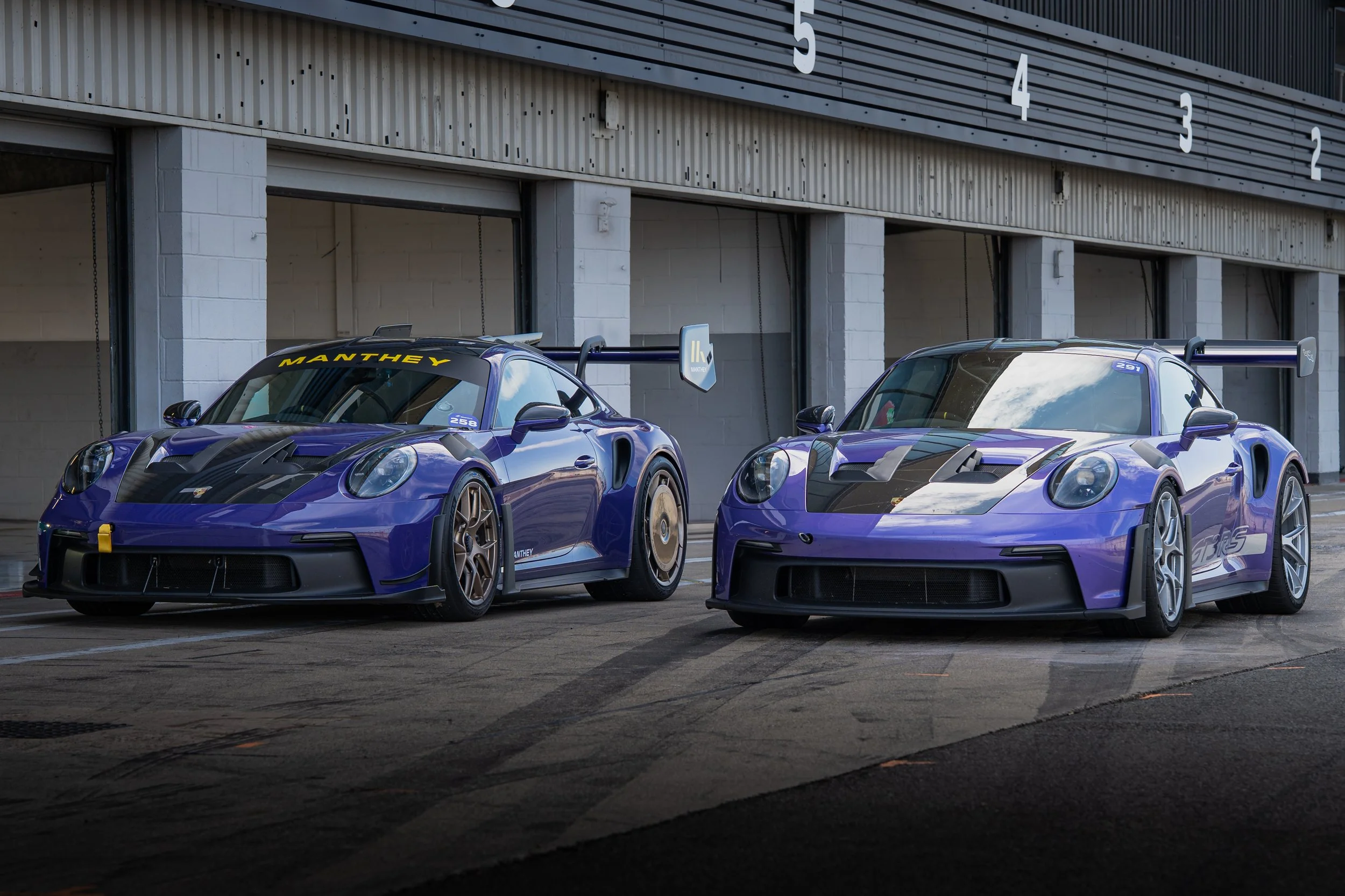 Two purple race cars parked side by side in a garage with the number 5 on the left car and the number 2 on the right car.