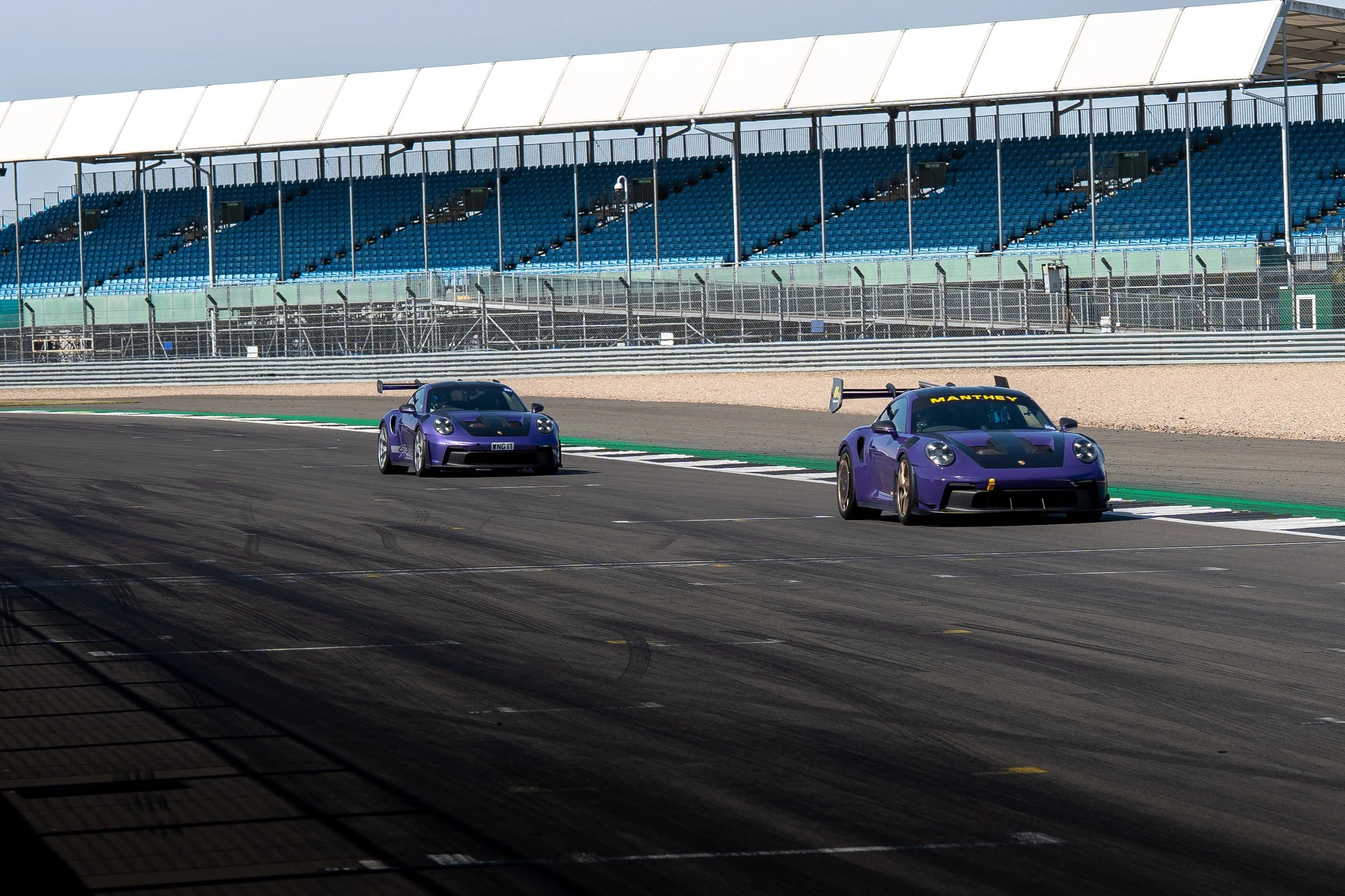 Two purple race cars driving on a race track with empty grandstands in the background.