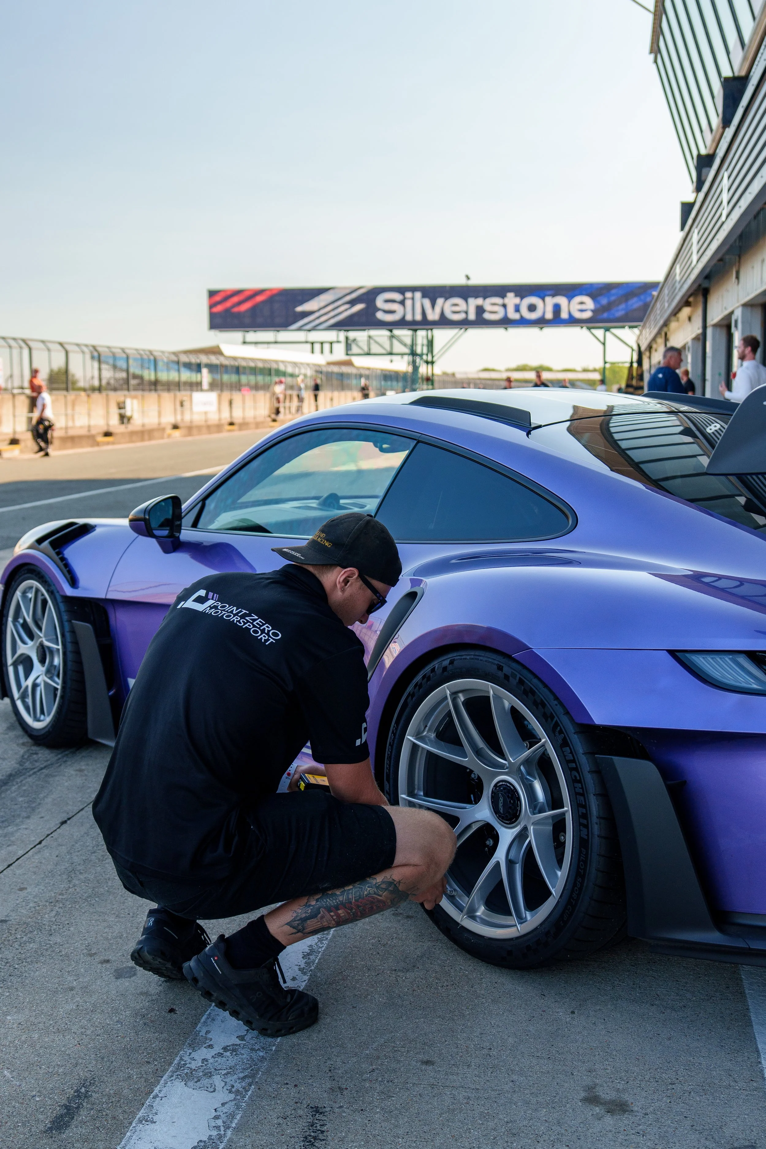 A man in a black T-shirt, shorts, and cap is fixing or inspecting a purple sports car in a pit lane at Silverstone race track. The car has racing tires and a spoiler, and is parked near a team garage with people talking in the background.