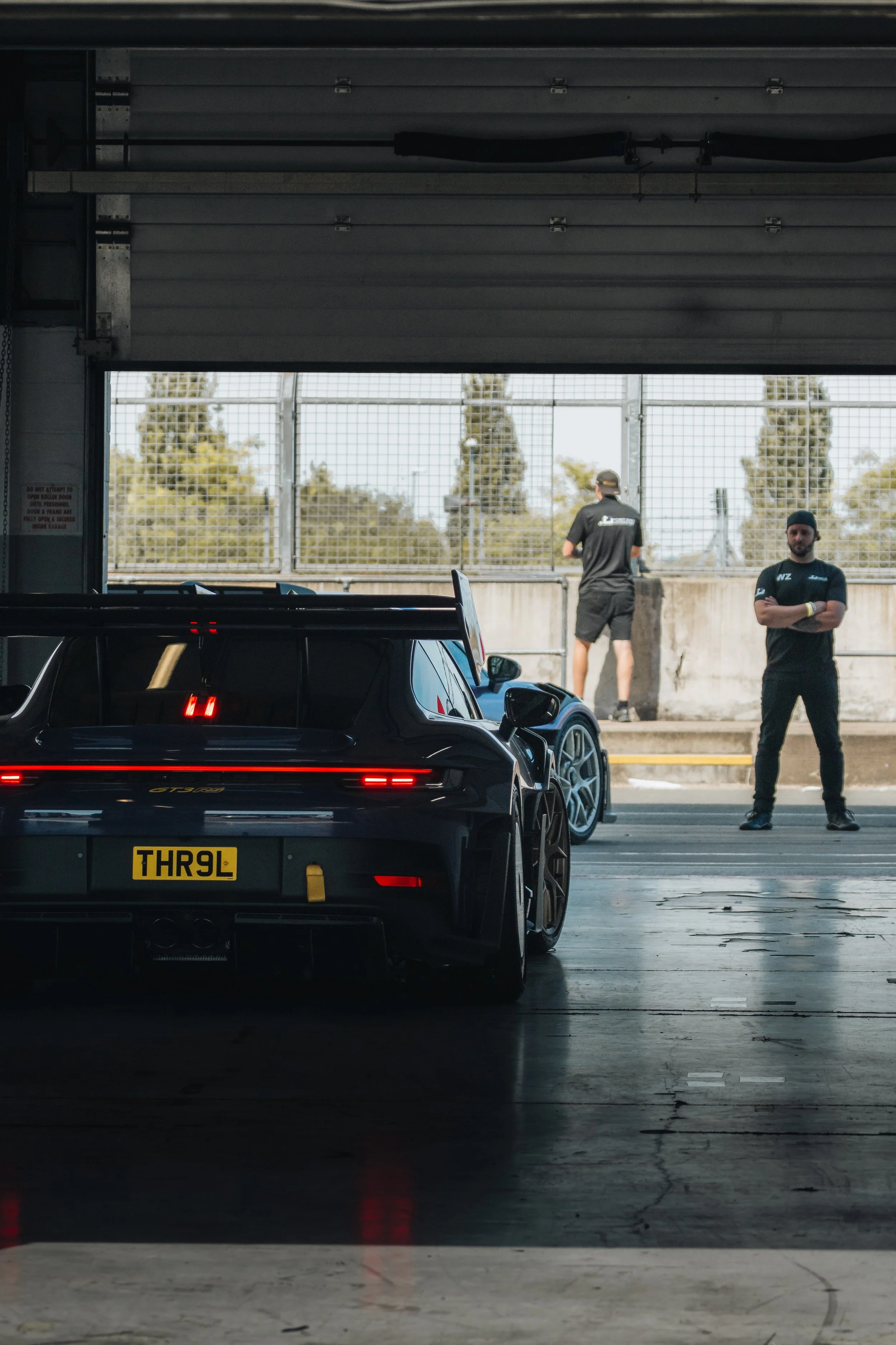 A black race car with a yellow license plate marked 'THR9L' inside a garage, with two individuals in black shirts outside near a fence, during daytime.