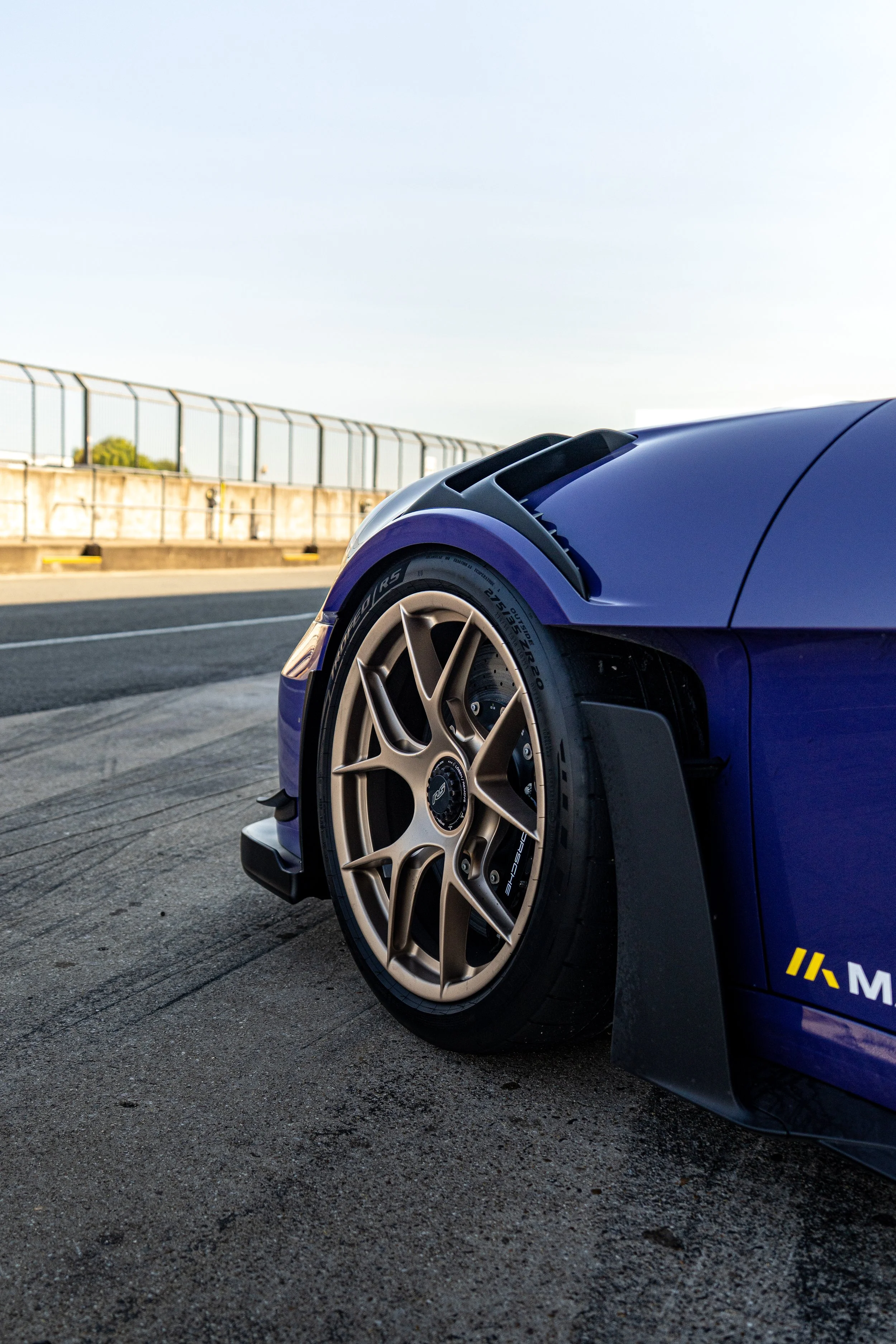 Close-up of a blue sports car with gold wheel rims, parked on a race track, showing part of the front tire and fender against a bright sky.