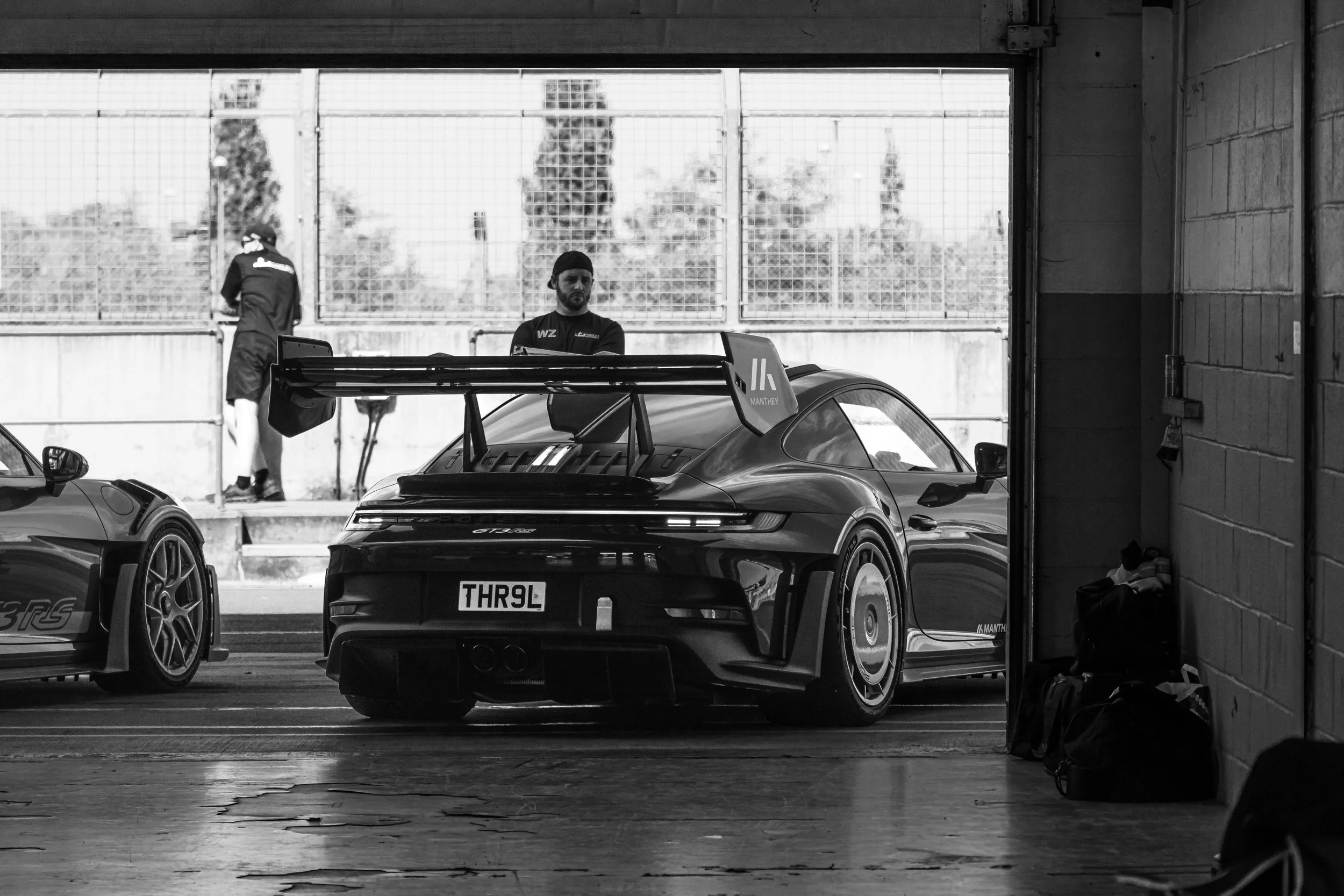 A black race car in a garage with two people in the background; one is standing and the other is looking out through a large open door, with trees visible outside.
