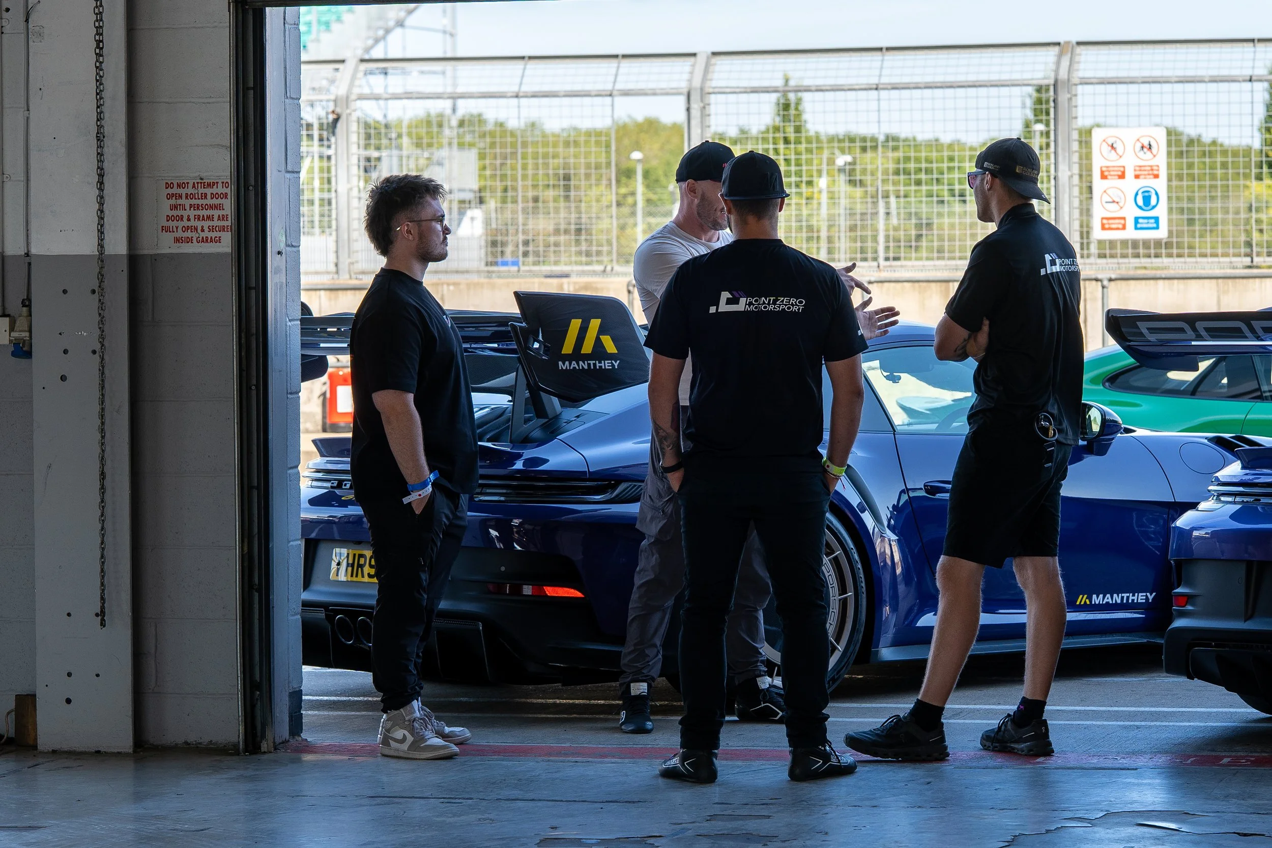 Four men standing and talking near a blue race car in a garage area with a fence and trees in the background.