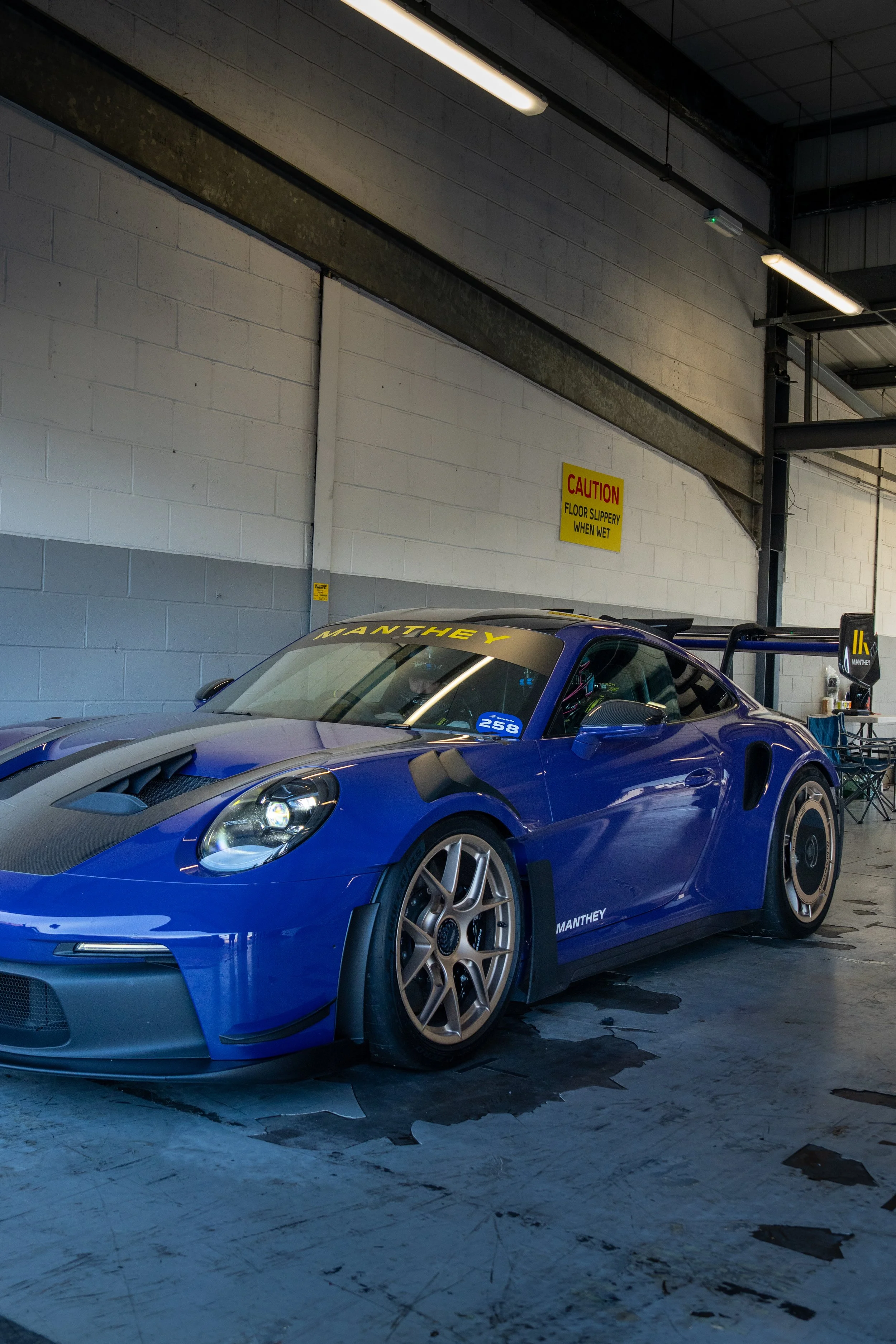 A race car with a blue body and gold wheels parked inside a garage, with a yellow 'Caution' sign on the wall behind it.