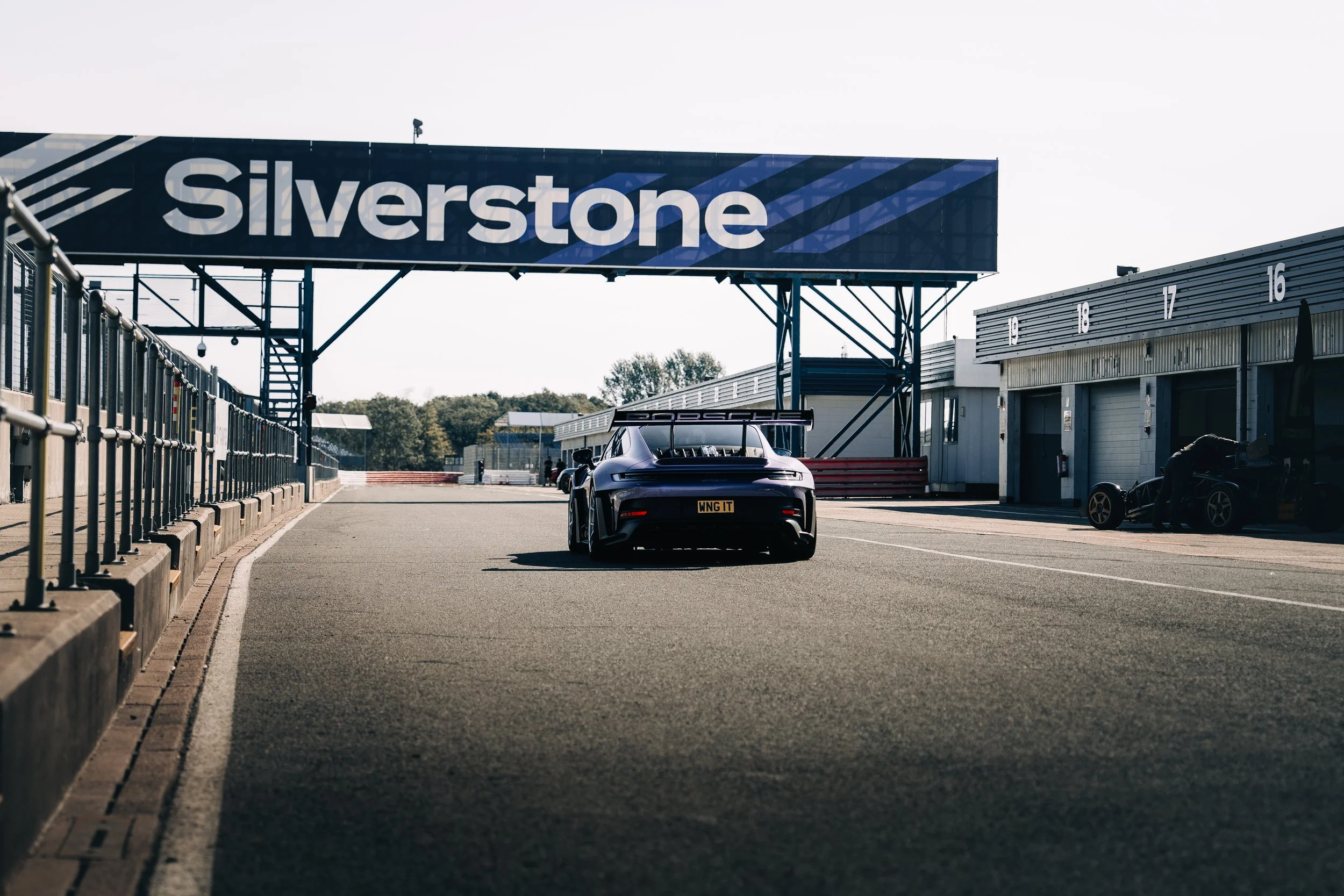 A race car on a track under a Silverstone sign with a garage and pit lane in the background.