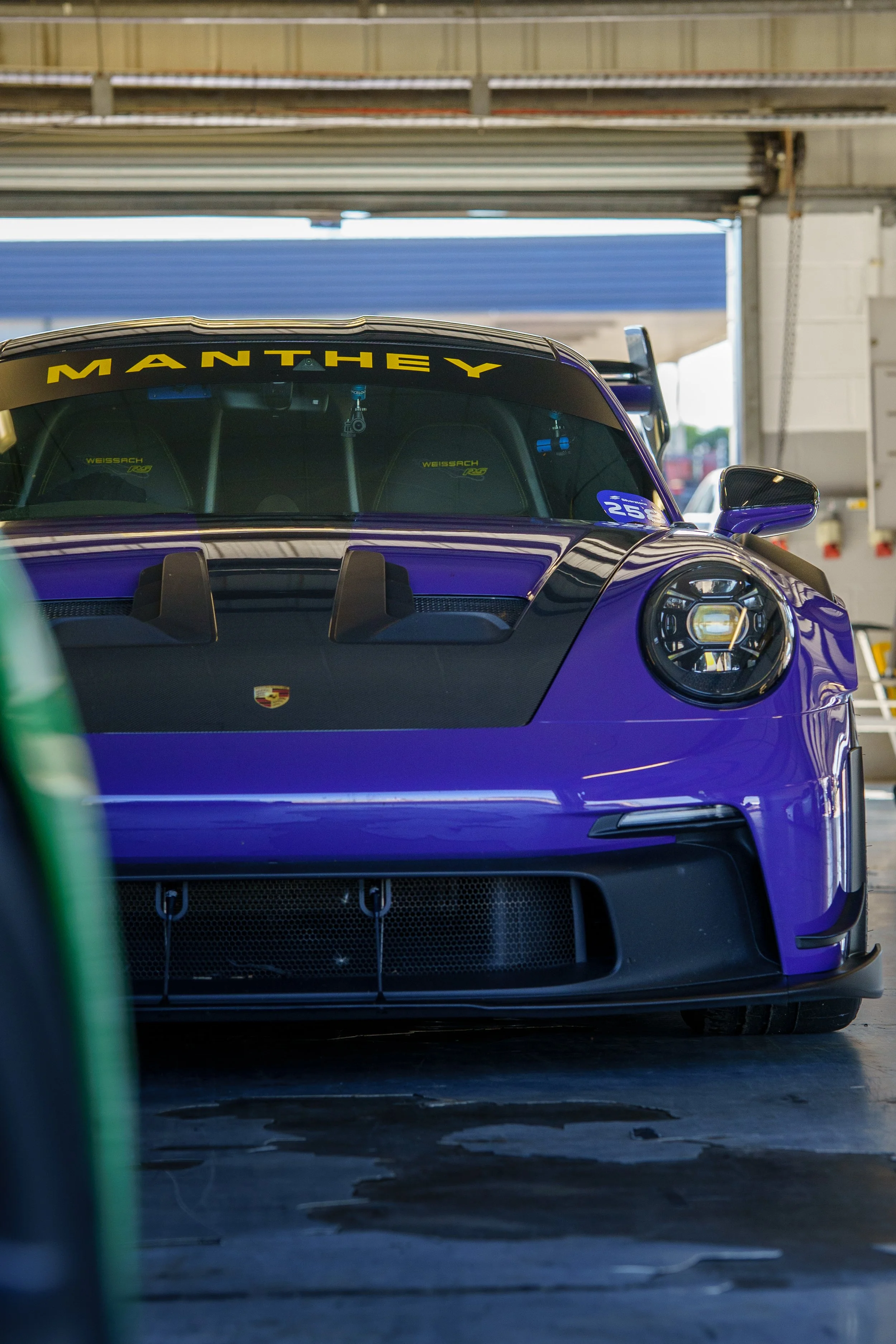 A purple Porsche race car inside a garage, with the word 'MANTHEY' in yellow at the top of the windshield and a Porsche logo on the front hood.