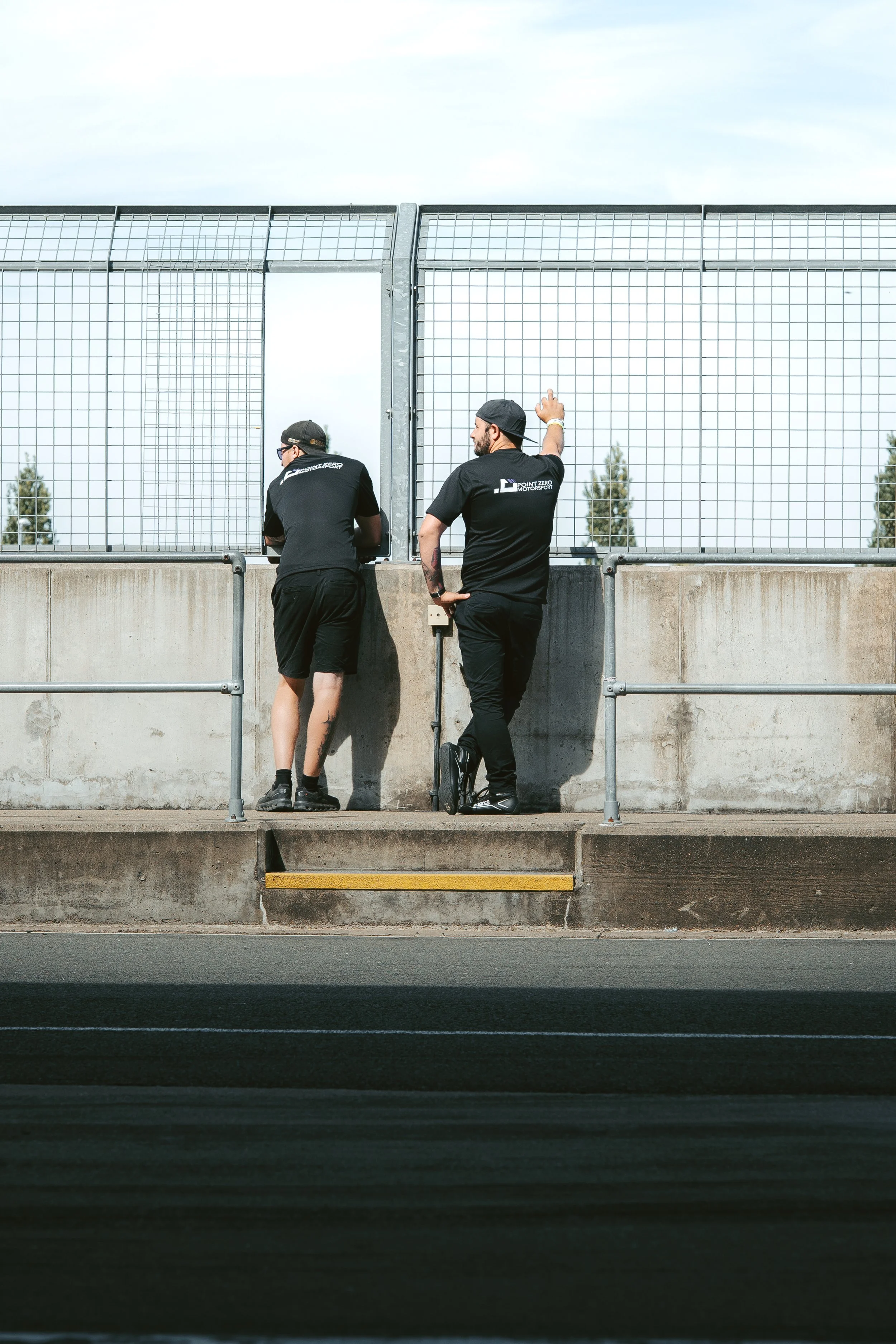 Two men in black clothing standing on a concrete barrier, looking through a chain-link fence at a race track.
