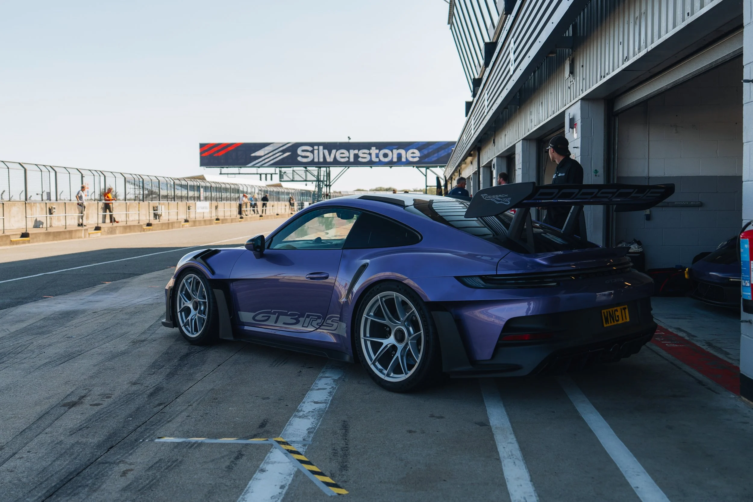 A purple Porsche GT3 RS race car parked in a pit lane at the Silverstone racing circuit, with team members preparing in the background.