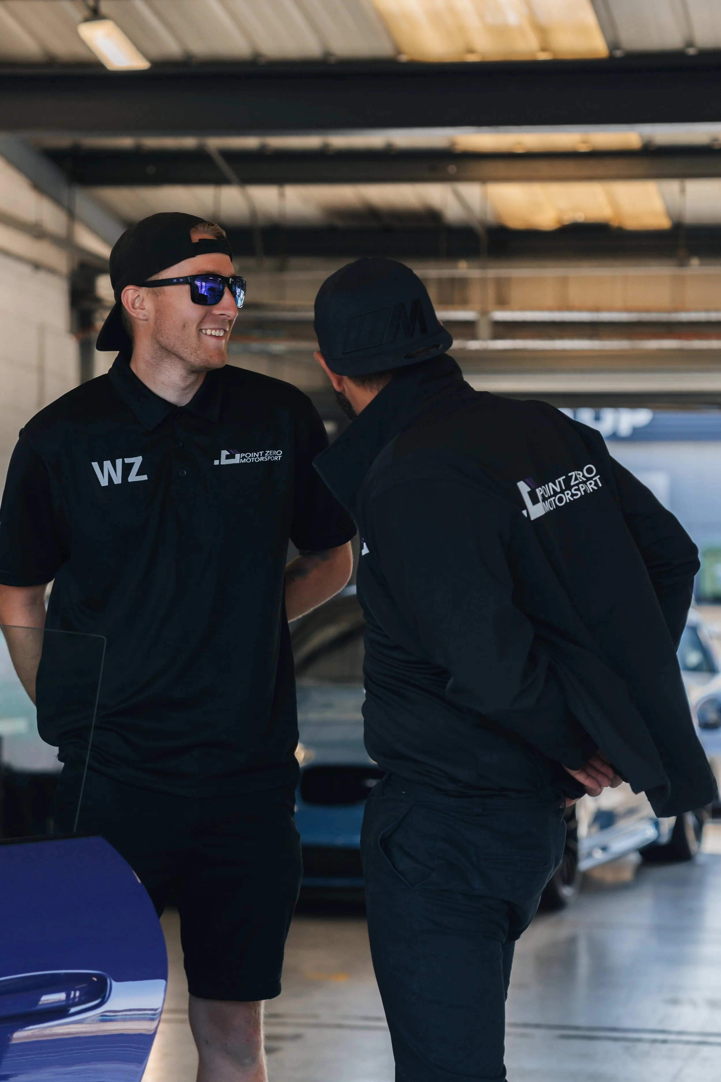 Two men wearing black Point Zero Motorsport shirts, smiling and conversing in a garage with race cars and a ceiling with exposed beams.