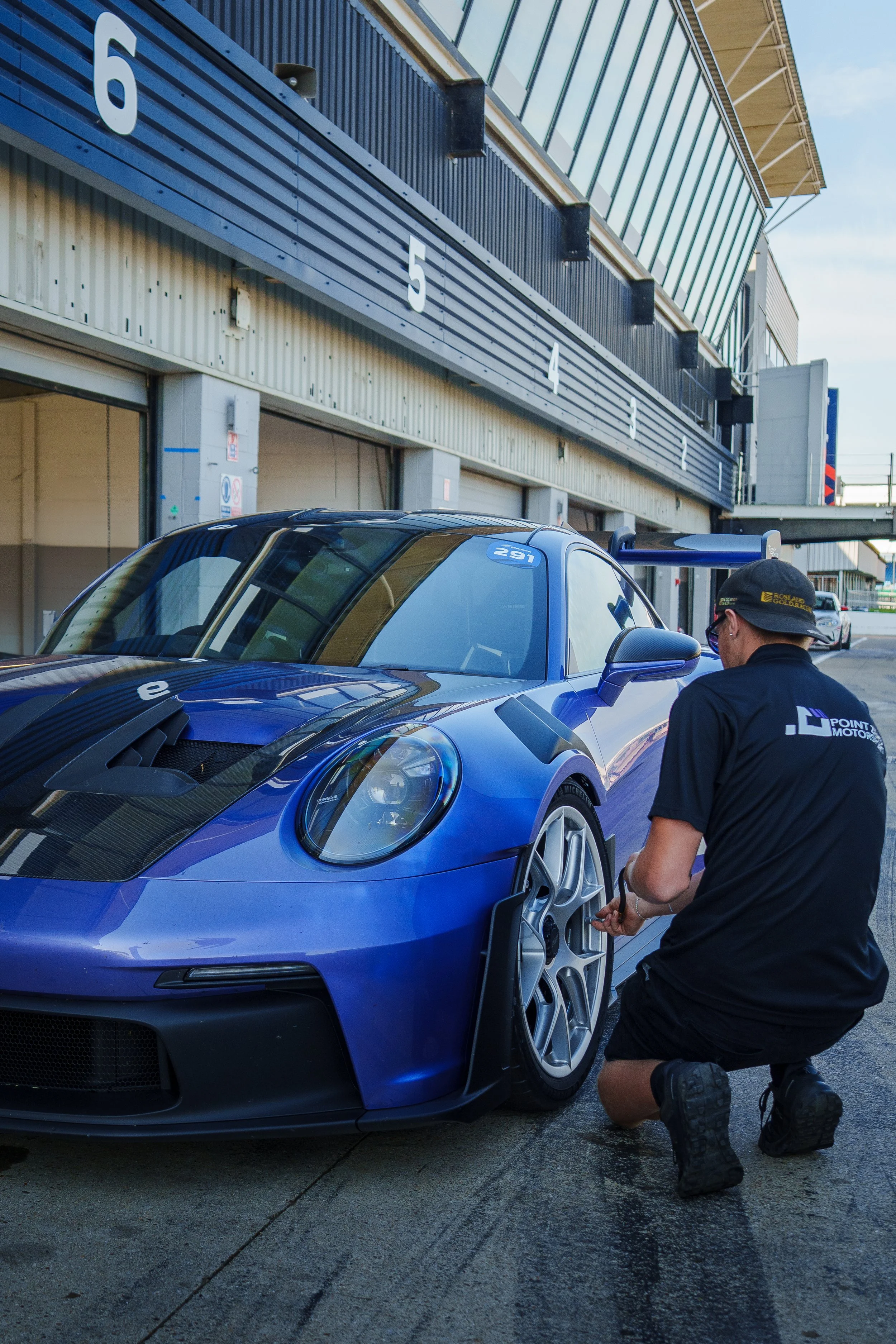 A person kneeling next to a blue race car, working on the front right tire at a racing track with garages in the background.