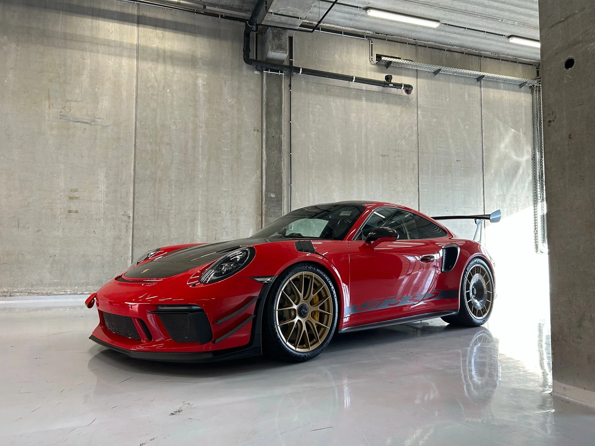 A red sports car with black accents parked in an industrial-style garage with concrete walls and a reflective white floor.