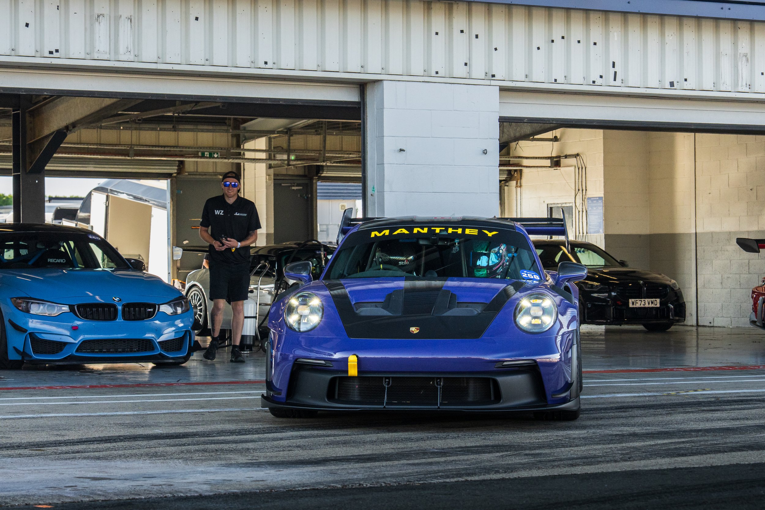 A purple racing car with yellow accents in a garage area, with a man in black clothing and sunglasses standing nearby, and other cars in the background.