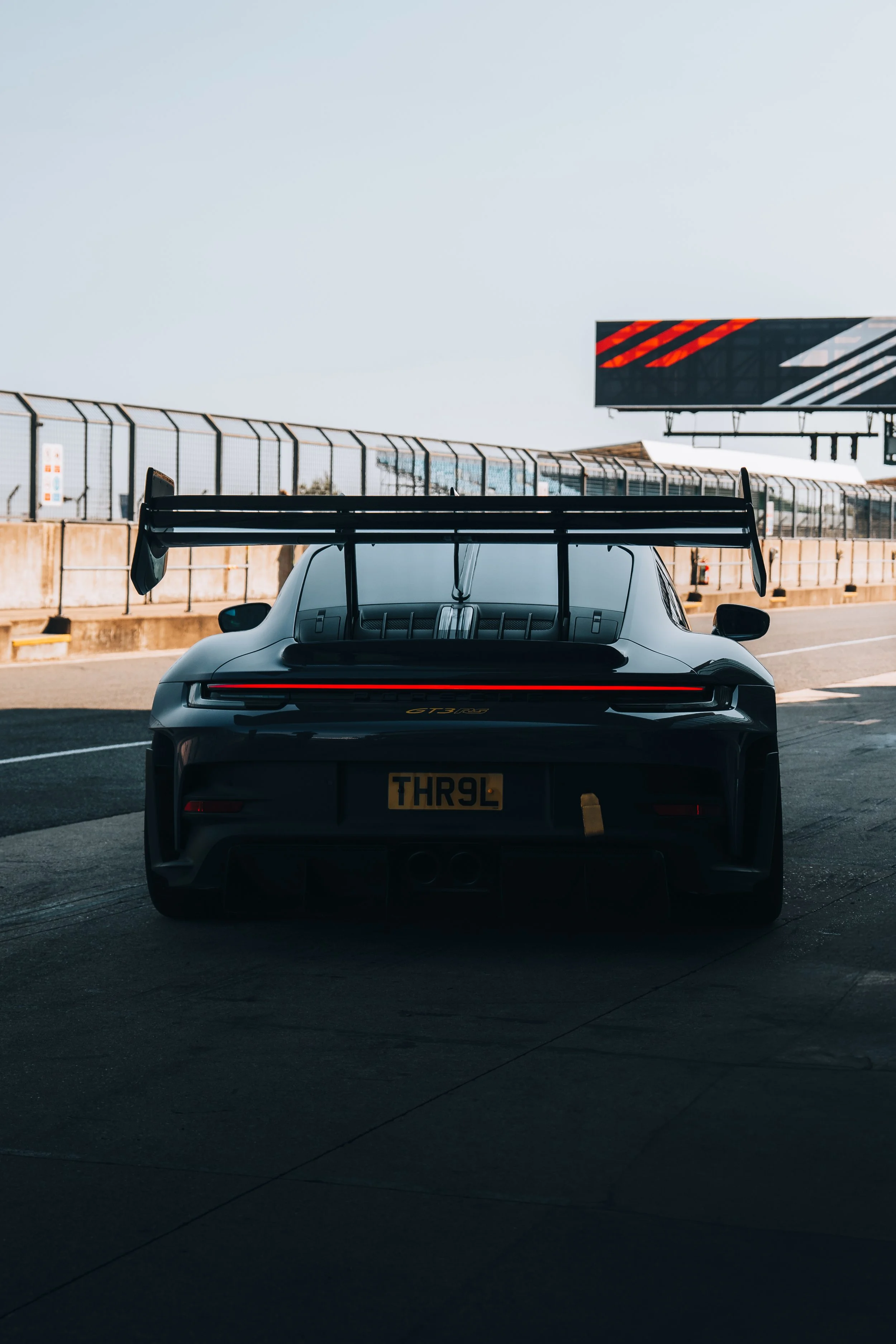 A black Porsche 911 GT3 race car on a race track, viewed from the rear with a large rear wing, grid markings on the track, a fence, and a billboard in the background.