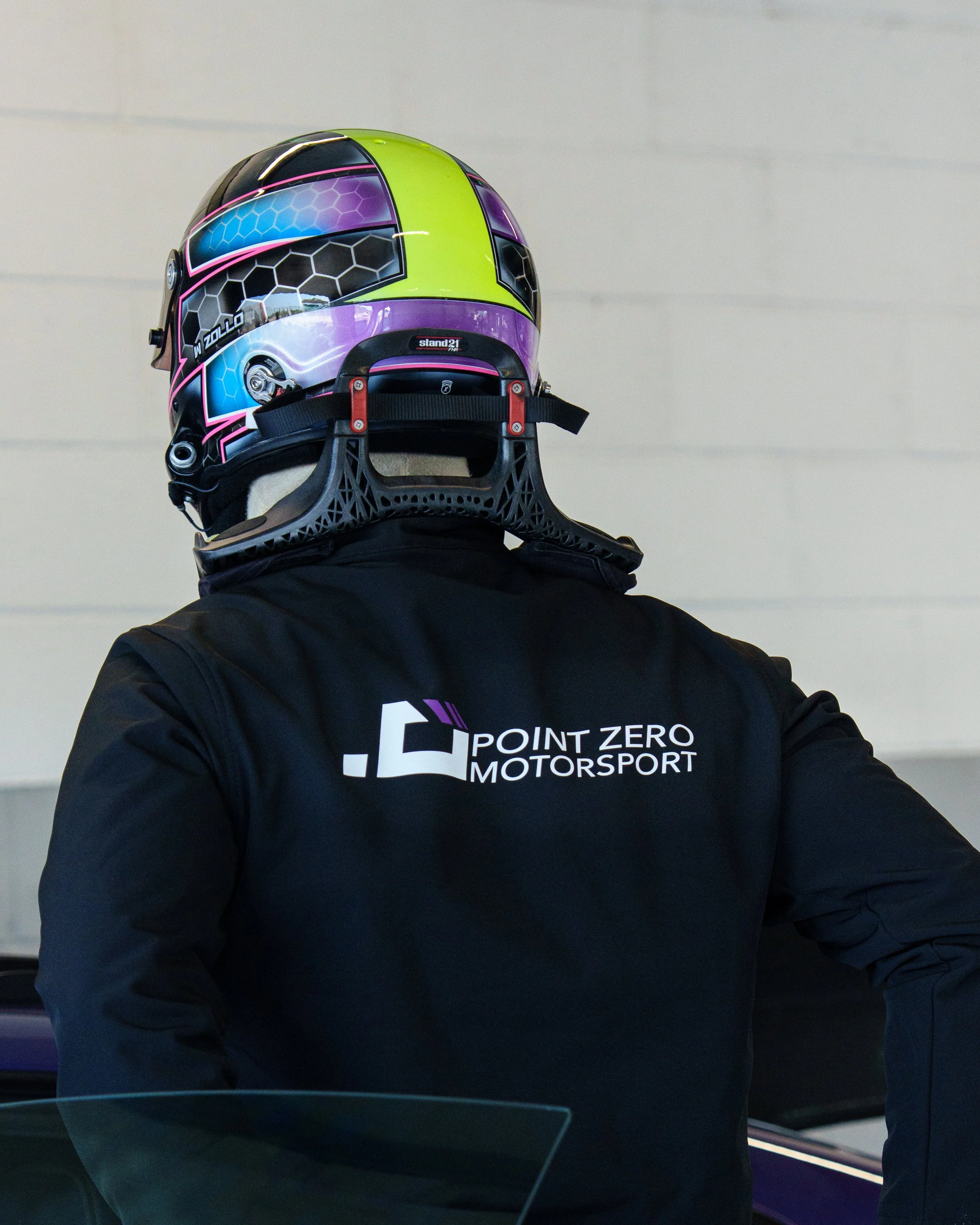 Person wearing a black jacket with 'Point Zero Motorsport' logo and a colorful racing helmet.