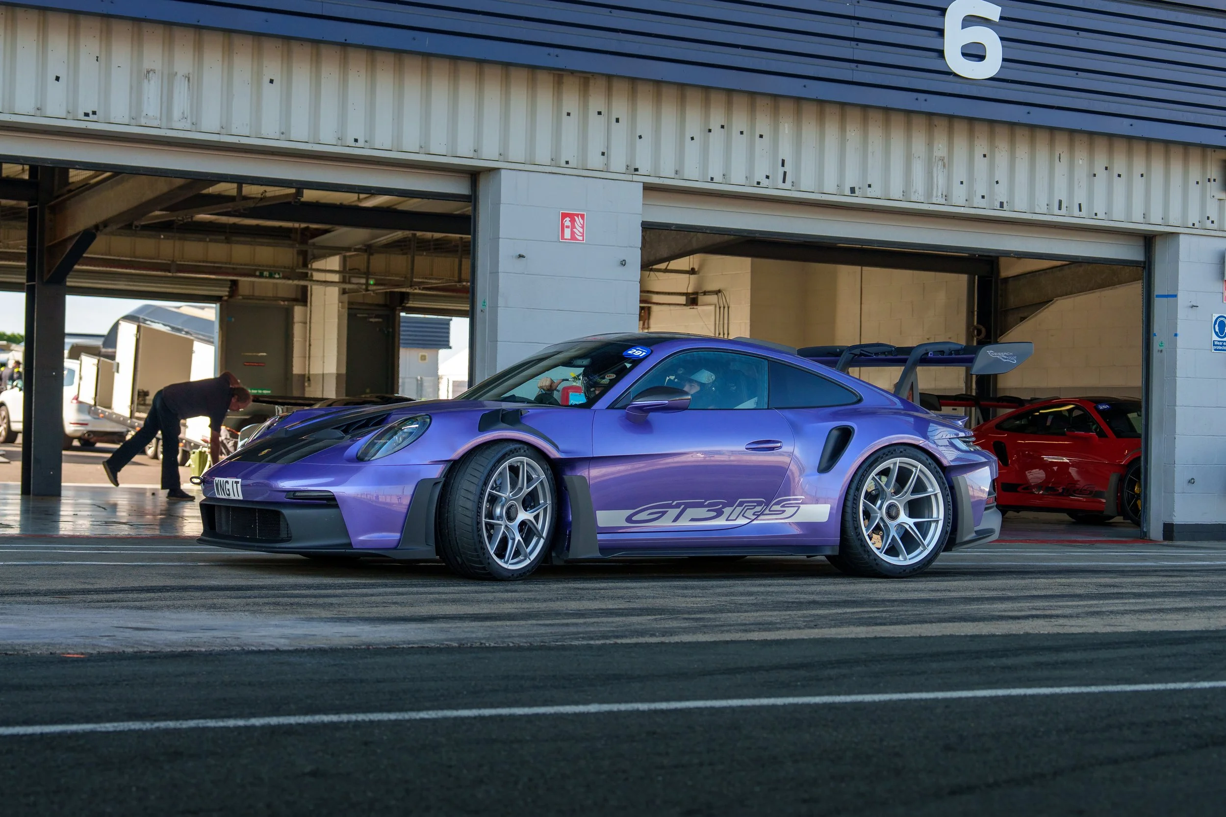 Purple Porsche 911 GT3 RS race car in a garage at a race track with a red car in the background.