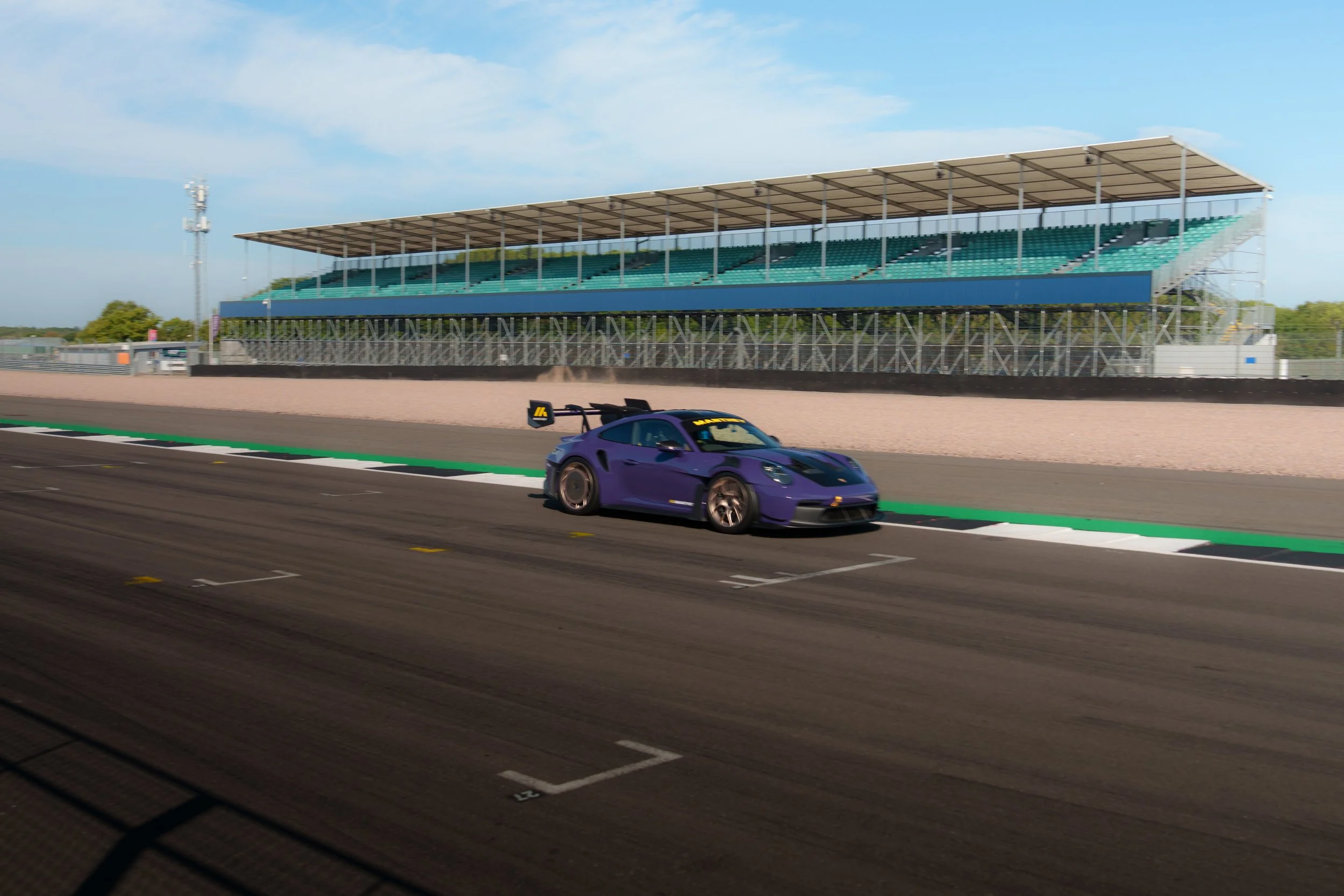 A purple sports car on a race track with a grandstand and clear blue sky in the background.