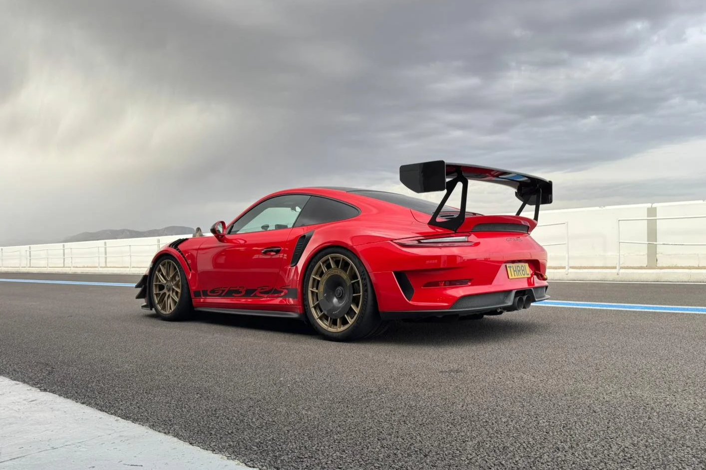 Red Porsche 911 GT3 RS with a large rear wing parked on a racetrack with dark cloudy skies in the background.