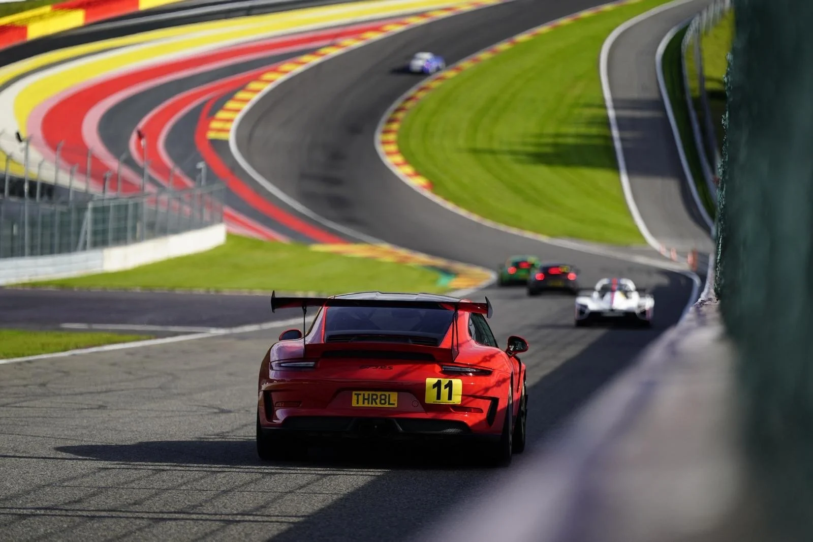 A red race car with the number 11 on the back drives on a winding race track with other race cars in the background, surrounded by green grass and colorful barriers.