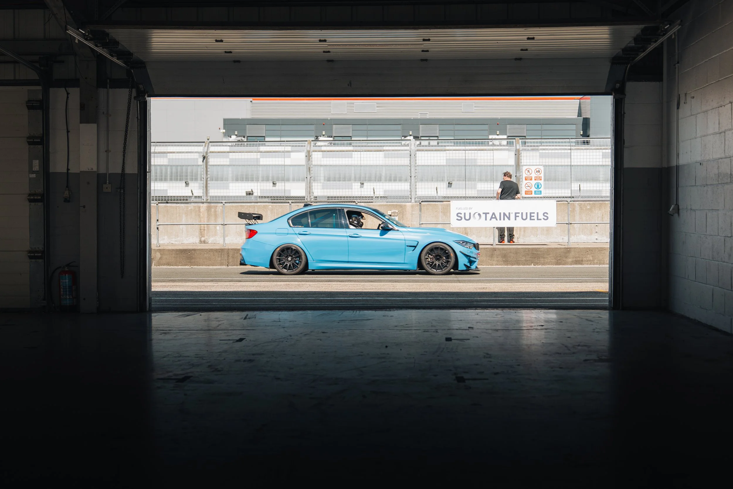 A blue race car with a large rear wing is parked on a race track, viewed from inside a dark garage, with a person standing behind a barrier holding a banner that reads 'Fueled by SU TAIN FUELS'.