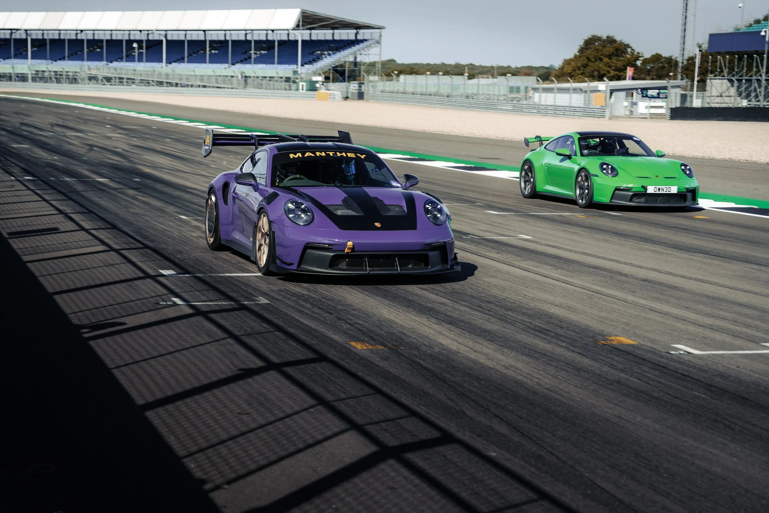 Two race cars, a purple Porsche with a black stripe and a green Porsche, are racing on a track.