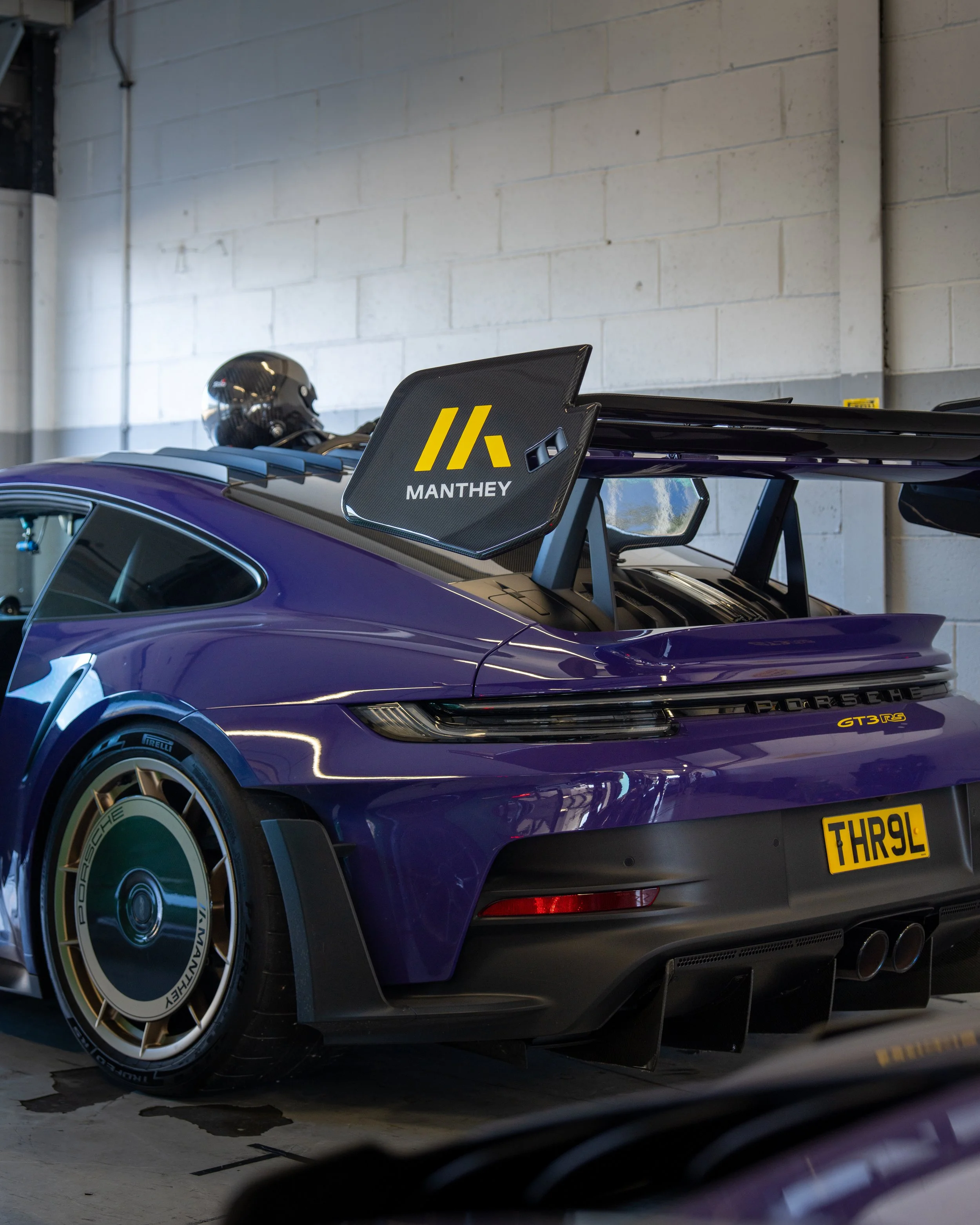 A purple Porsche 911 GT3 RS sports car with racing decals and a large rear wing, parked indoors near a brick wall, with a racing helmet on the roof.