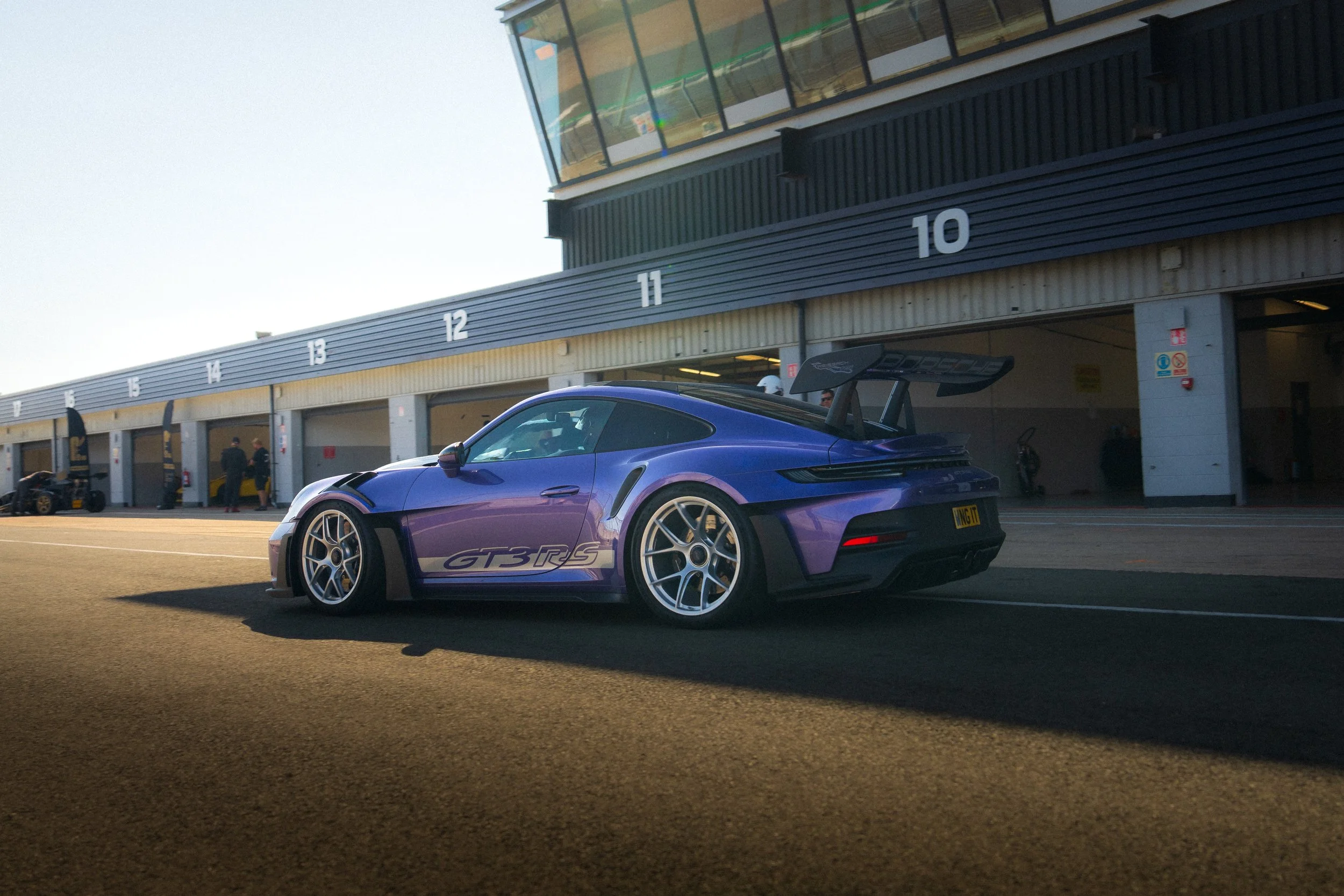 A purple Porsche 911 GT3 RS parked at a racetrack pit lane with garages numbered 10 to 15 in the background.