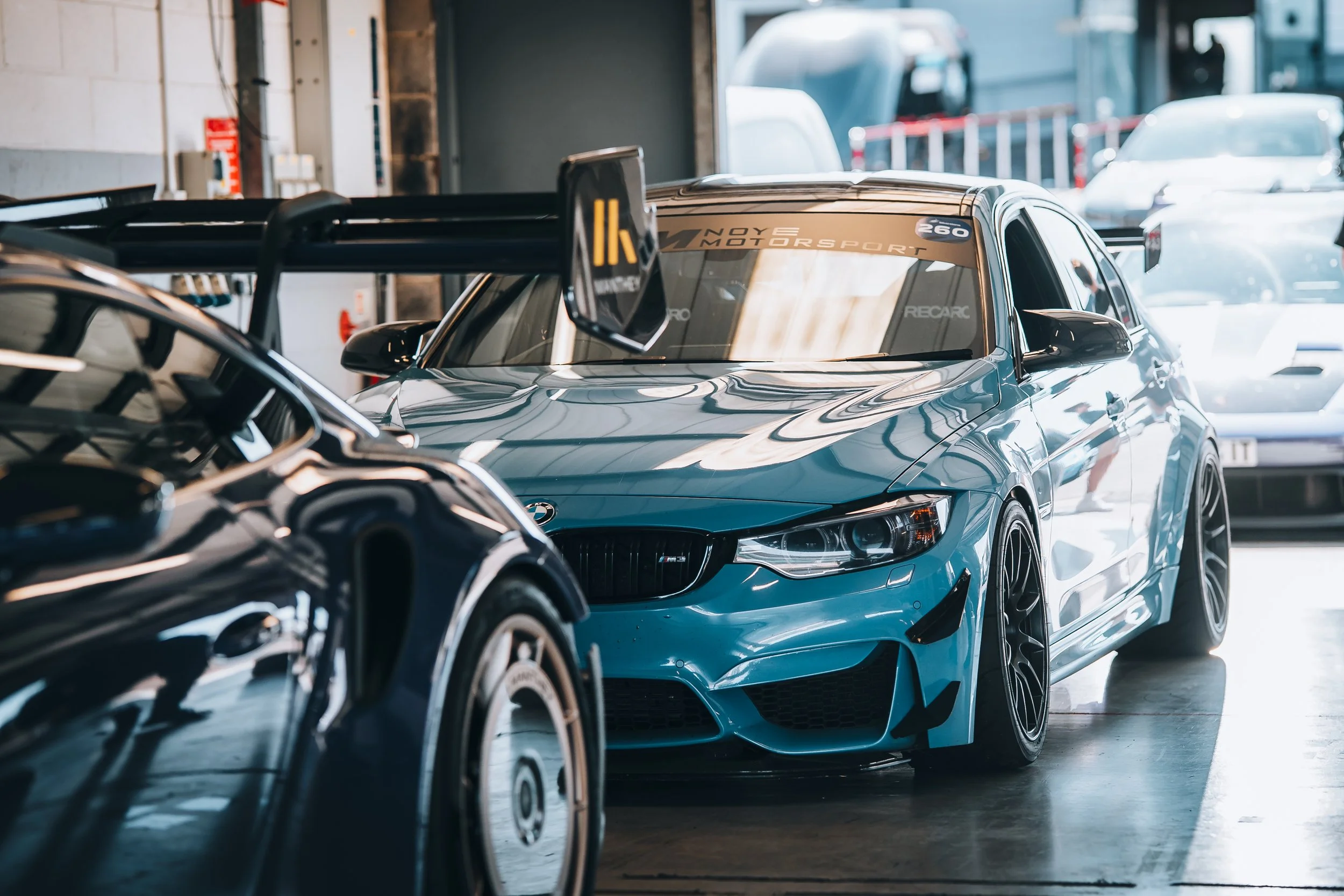 A blue BMW race car with carbon fiber side mirrors and a large rear wing is parked in a garage, surrounded by other cars.