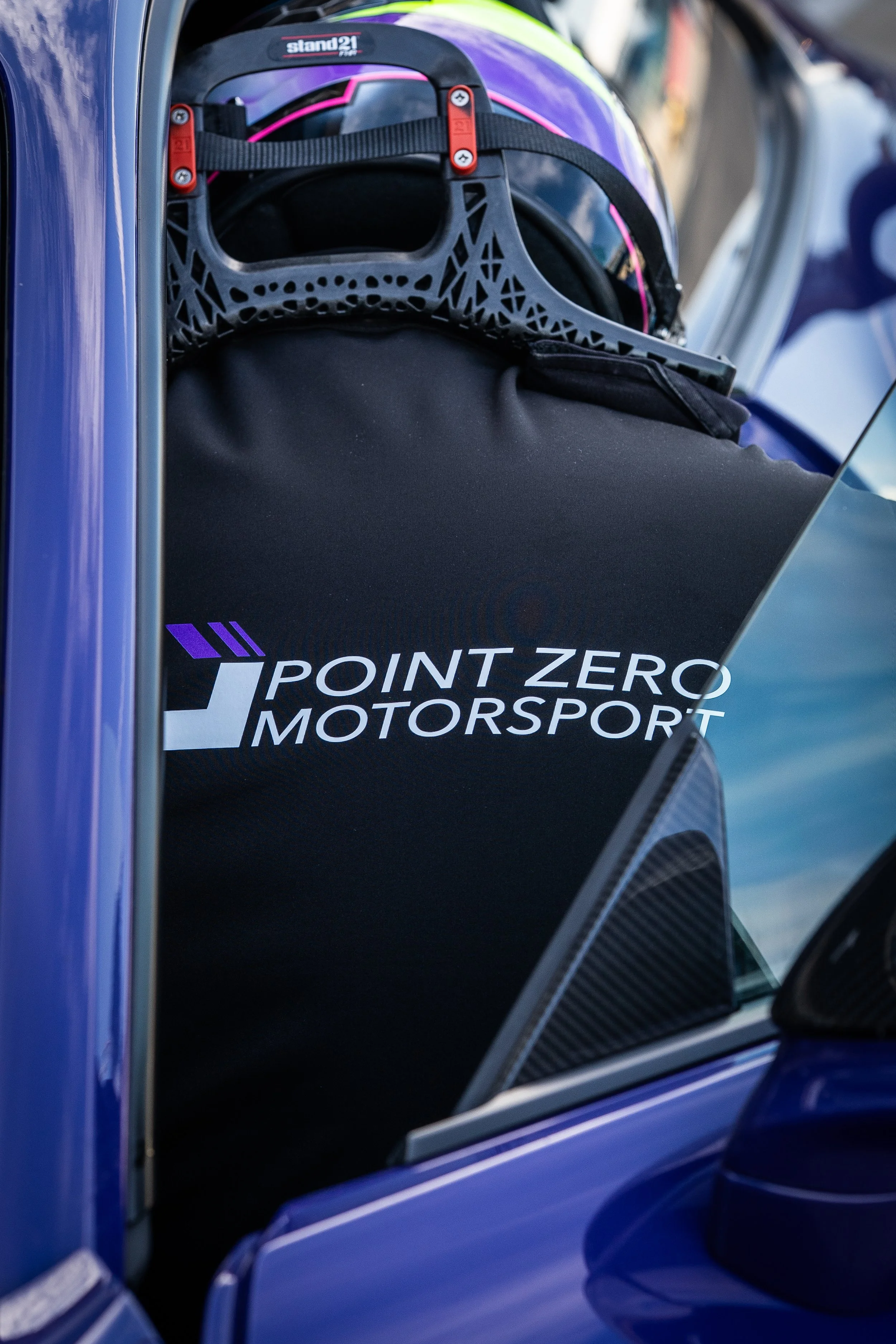 View of a helmet inside a racing car with a black fabric cover displaying the Point Zero Motorsport logo, seen through the car window.