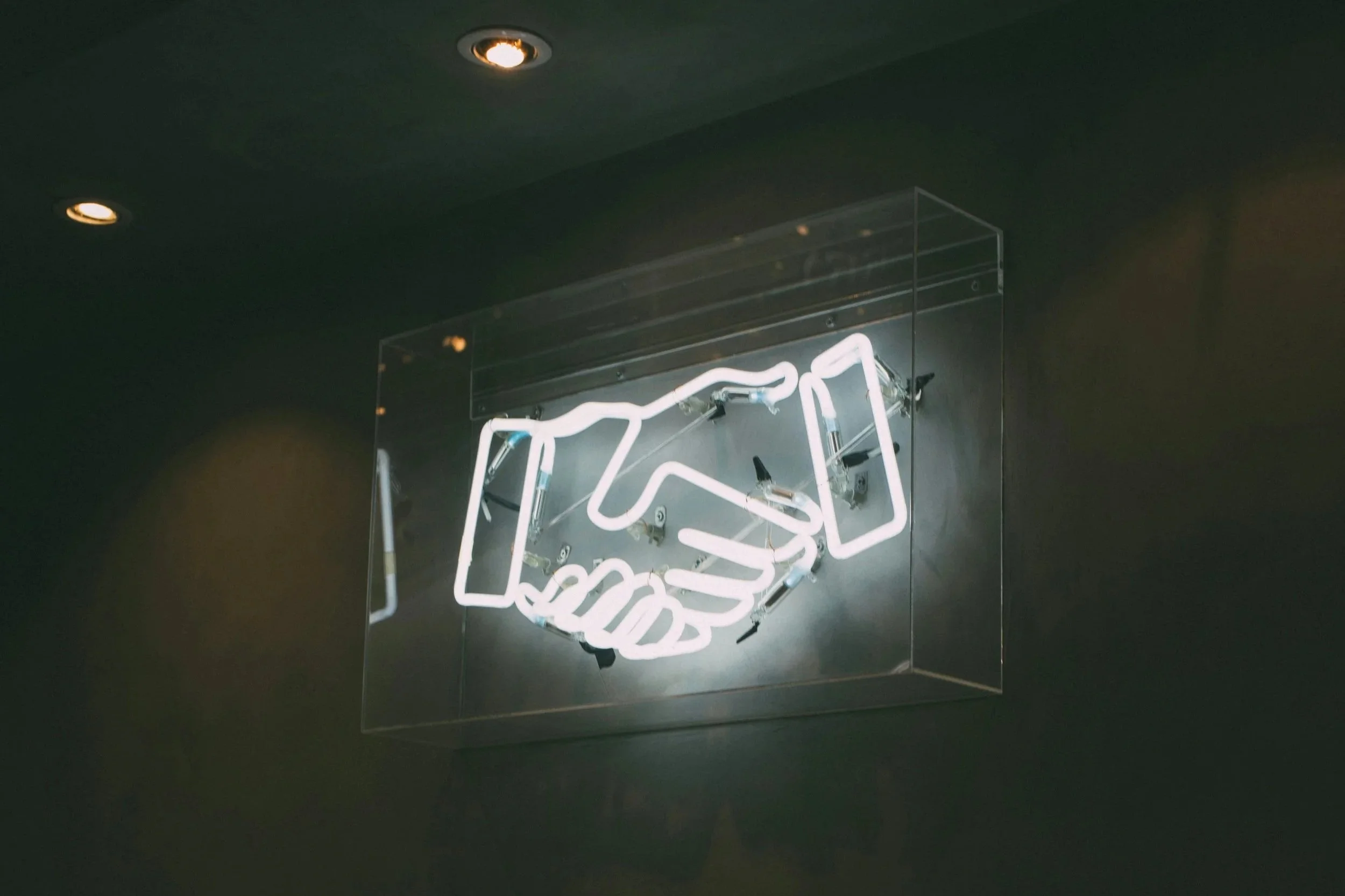 A white neon sign hung up on a wall underneath a wire bracket. The neon sign is of two hands shaking.