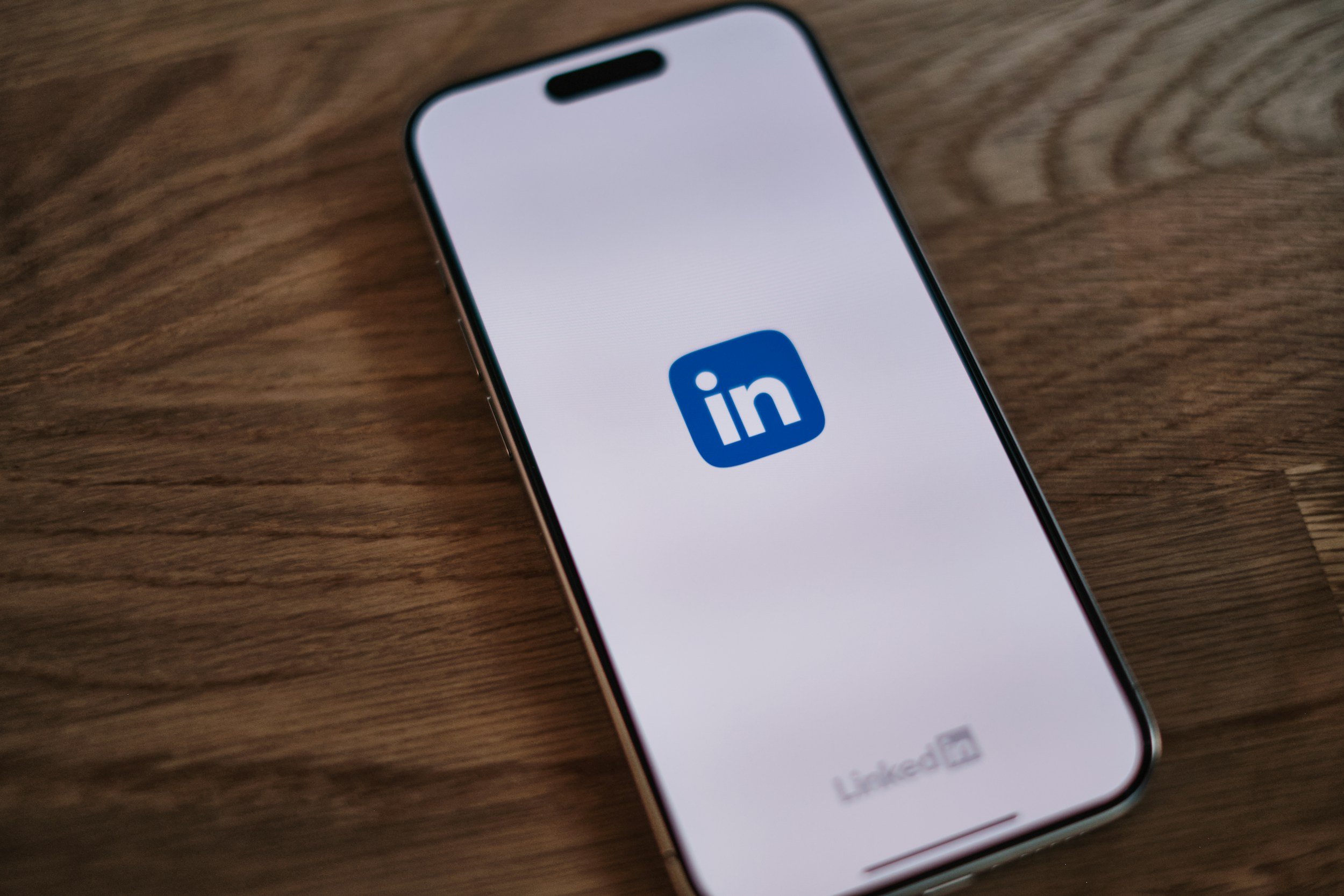 A mobile phone on a table, facing screen-up with the LinkedIn loading page on it. This a black white screen with a small, blue LinkedIn logo on.