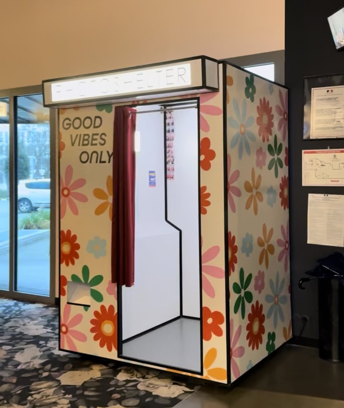 Cabine photo avec décoration de fleurs colorées, signe 'Photo Booth' en haut, écrit 'Good Vibes Only' sur le mur, rideau rouge devant l'entrée.