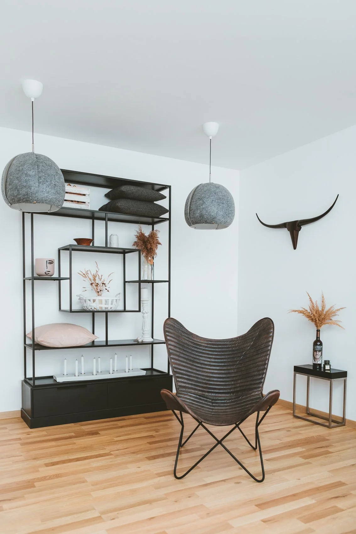 Moderne Wohnzimmergestaltung mit schwarzem Regal, Dekorationsgegenständen, einer Wandkunst mit Geweih und einem gemütlichen Sessel auf Holzboden.