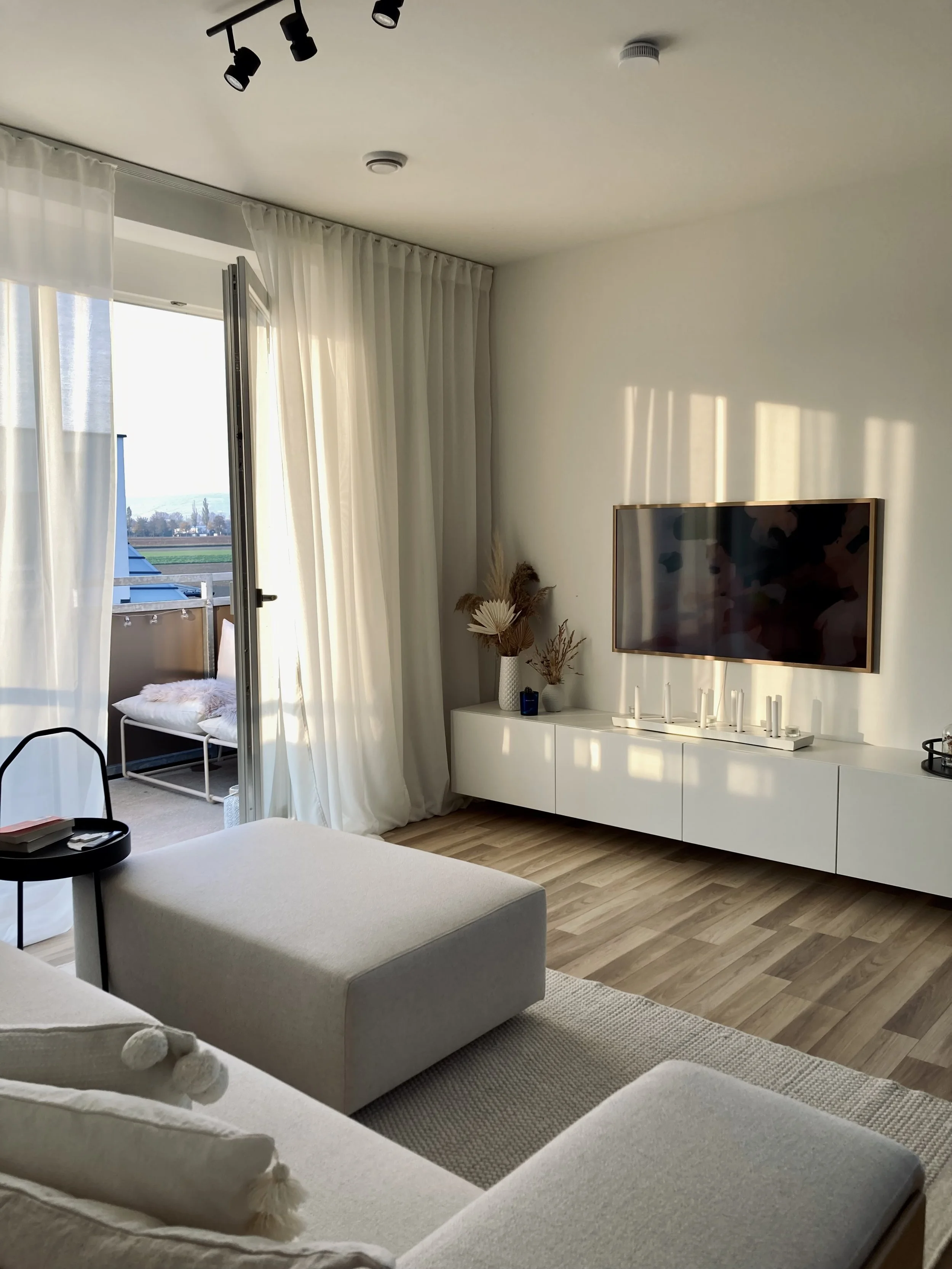 Ein modernes Wohnzimmer mit weißen Möbeln, einem flachbildschirm Fernseher an der Wand, Couchtisch, offene Balkontür mit Blick ins Freie, Vorhänge und dekorative Gegenstände wie Vasen mit Trockenblumen.